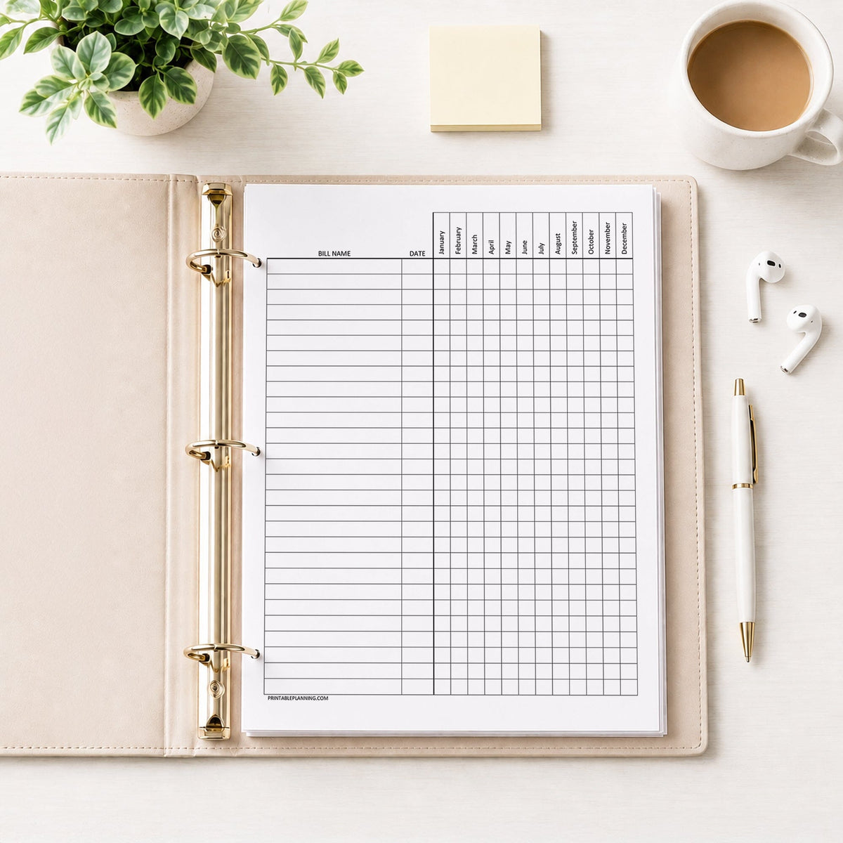 A printed Yearly Bill Payment Tracker - Printable Annual Bill Checklist (printable) page shown in a beige binder on a desk with a plant, sticky notes, coffee, earbuds, and a pen.