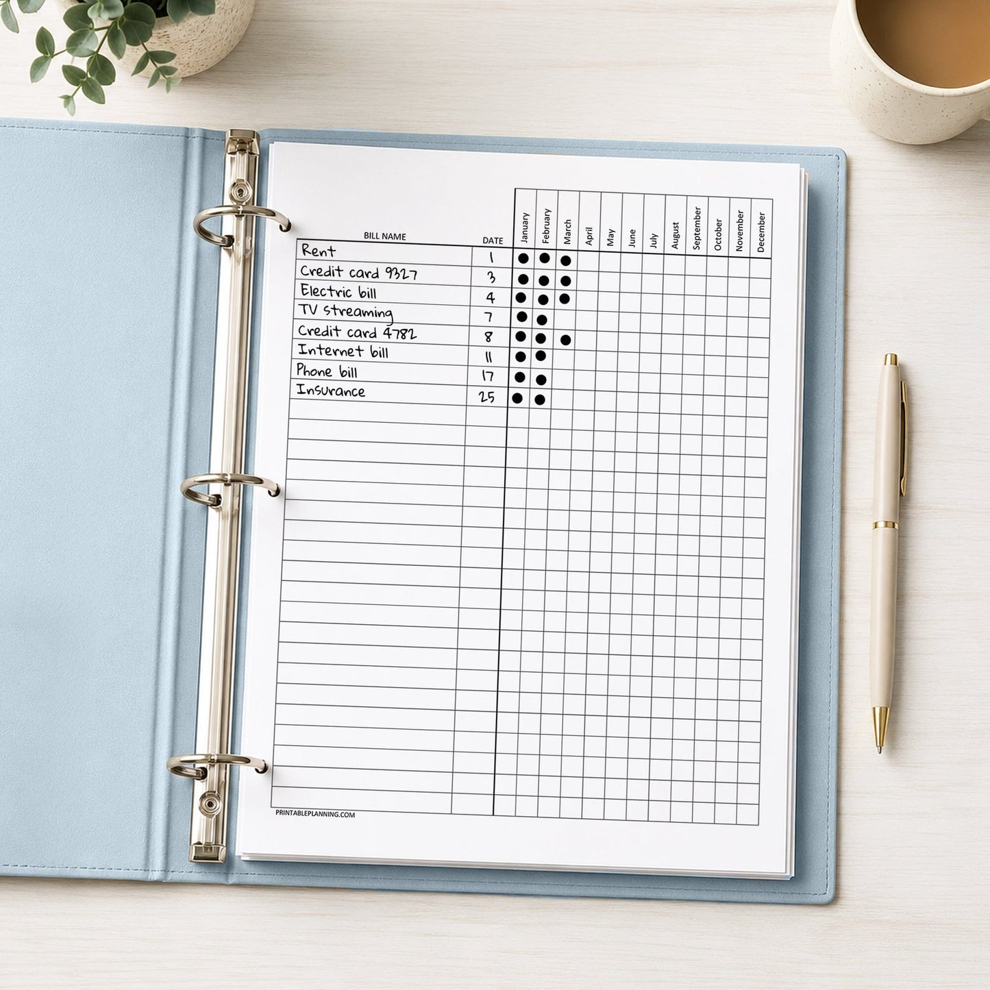 A printed Yearly Bill Payment Tracker - Printable Annual Bill Checklist (printable) page, shown with example writing, in a light blue binder on a desk with a plant, coffee, and a pen.