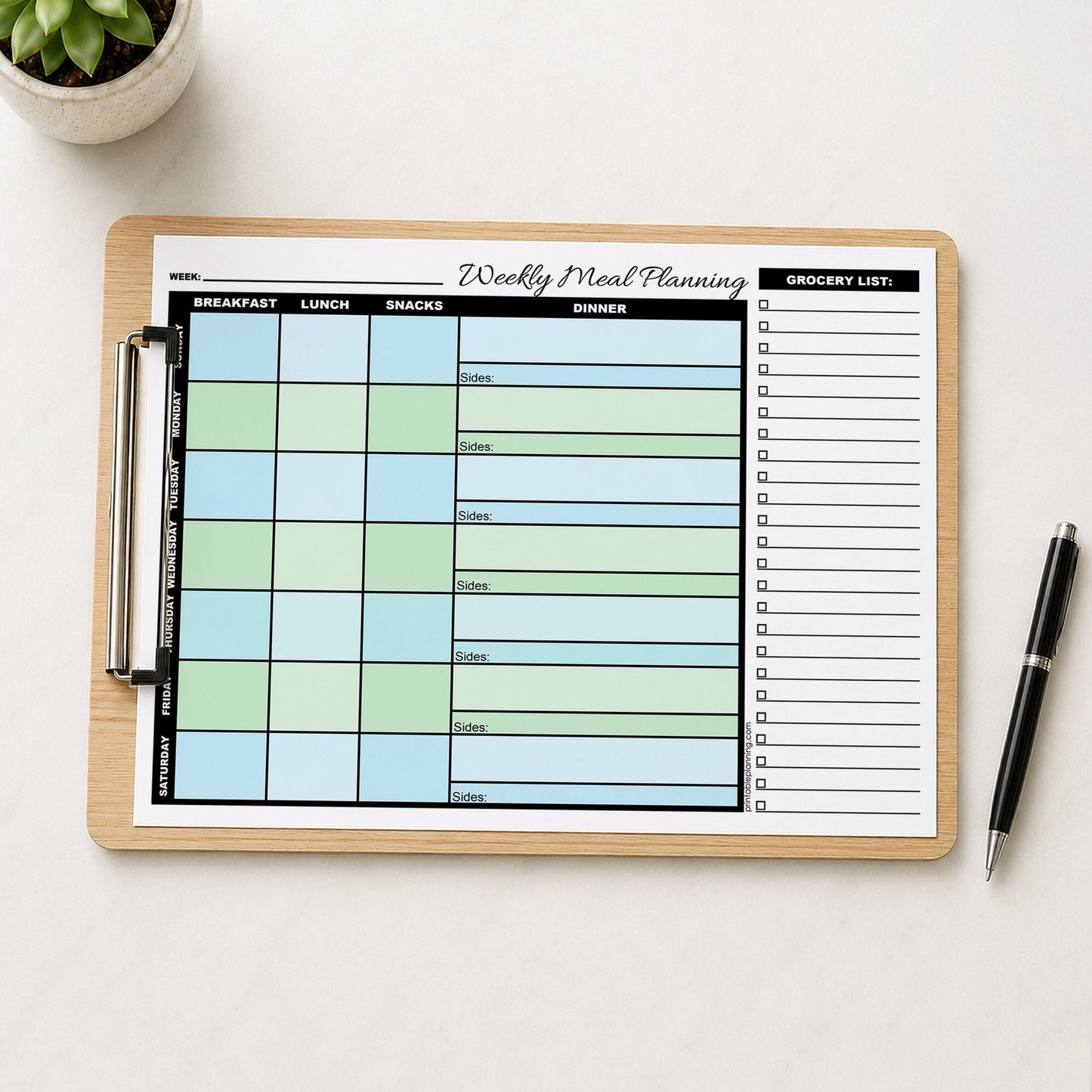 A printed Blue Green Weekly Meal Planner with Grocery List (printable page) shown on a wooden clipboard on a table with a plant and a pen.