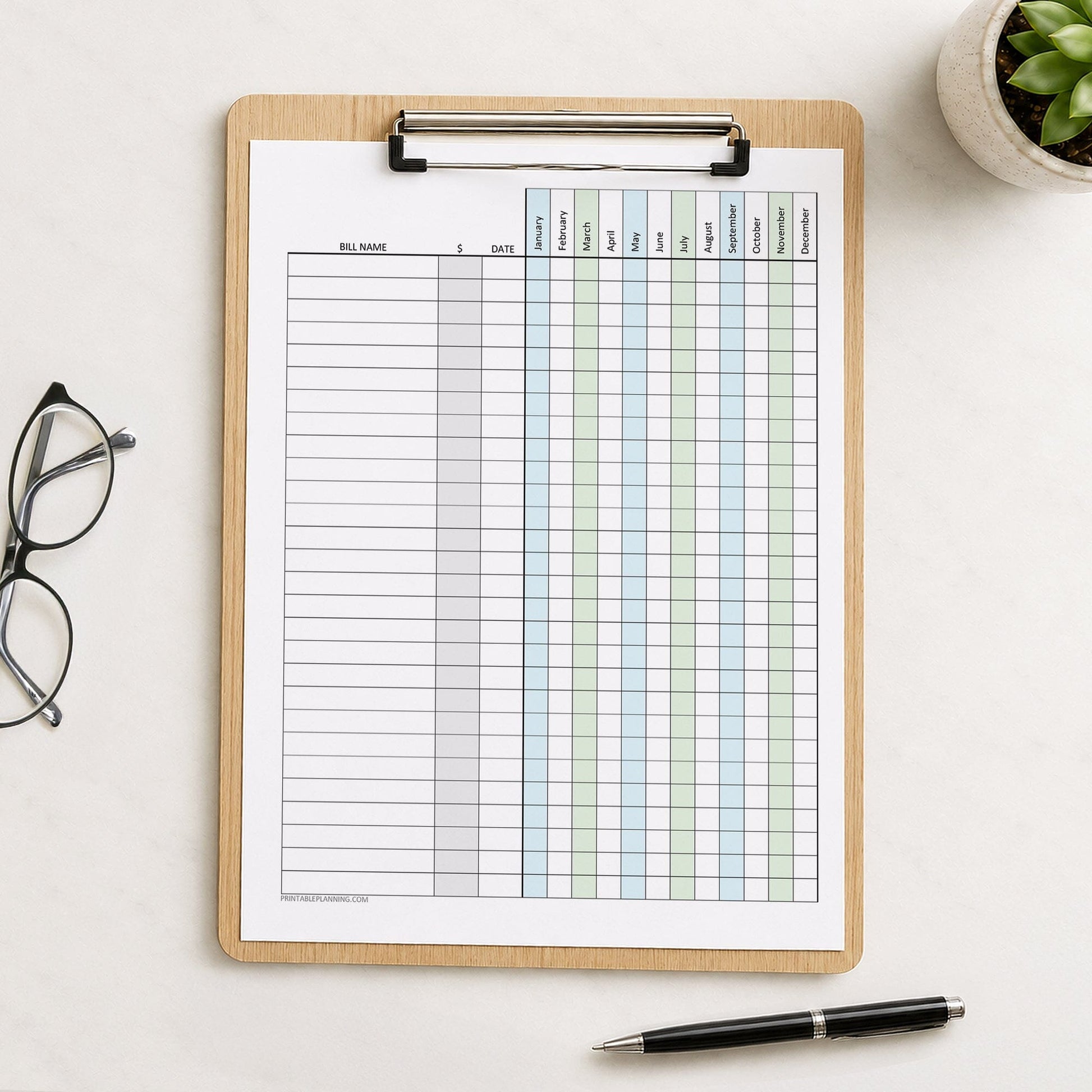 A wooden clipboard holding a Blue Green Yearly Bill Payment Tracker with Amount Column (printable page) styled on a light surface with eyeglasses, a small plant, and a pen nearby.