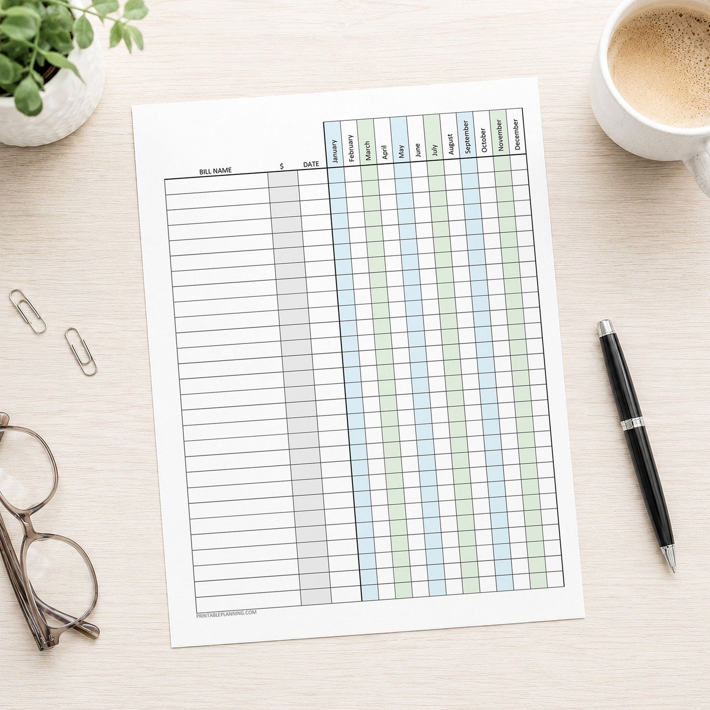 Blue Green Yearly Bill Payment Tracker with Amount Column (printable page) shown on a desk with glasses, a plant, paperclips, coffee, and a pen.
