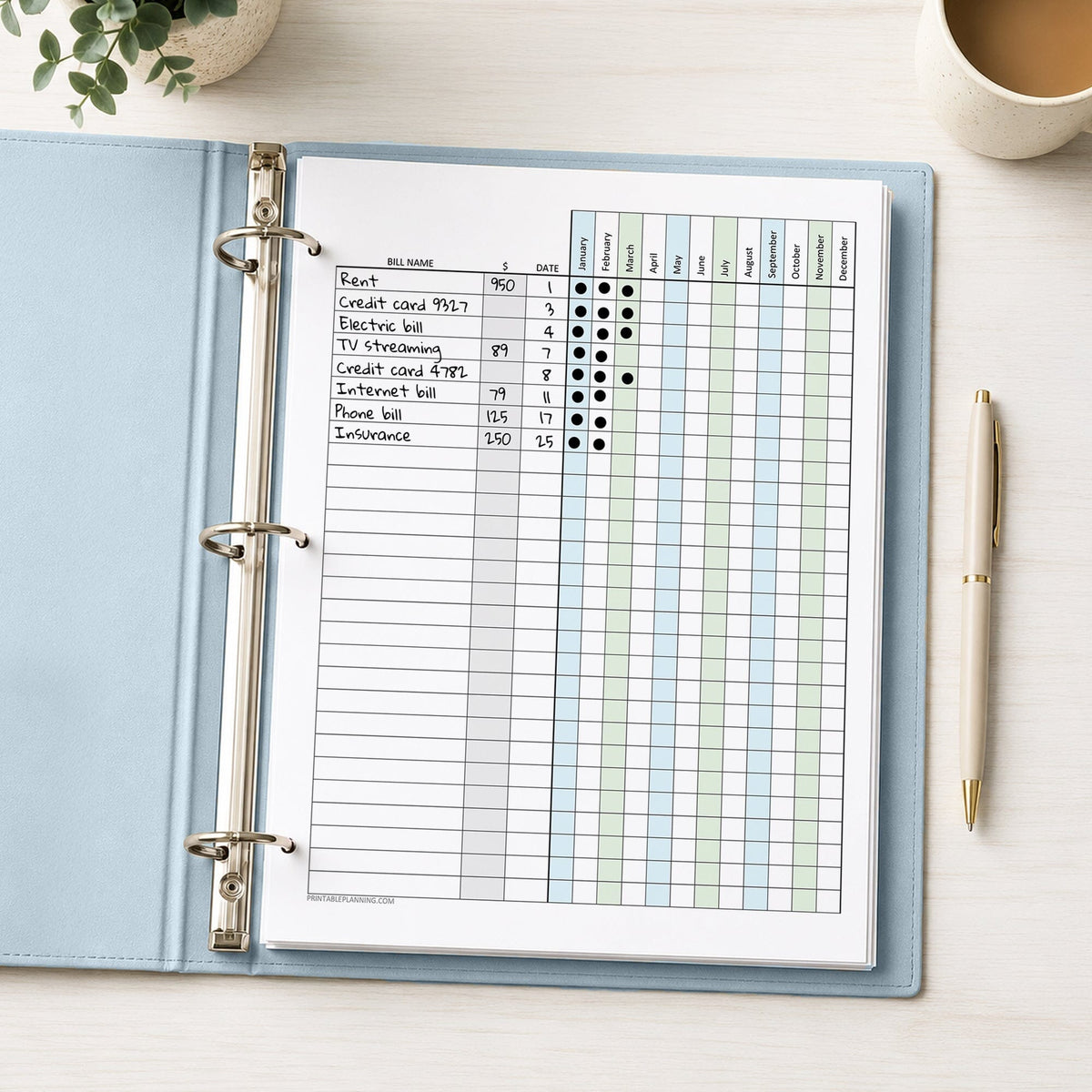 A printed Blue Green Yearly Bill Payment Tracker with Amount Column (printable) page, shown with example writing, in a light blue binder on a desk with a plant, coffee, and a pen.