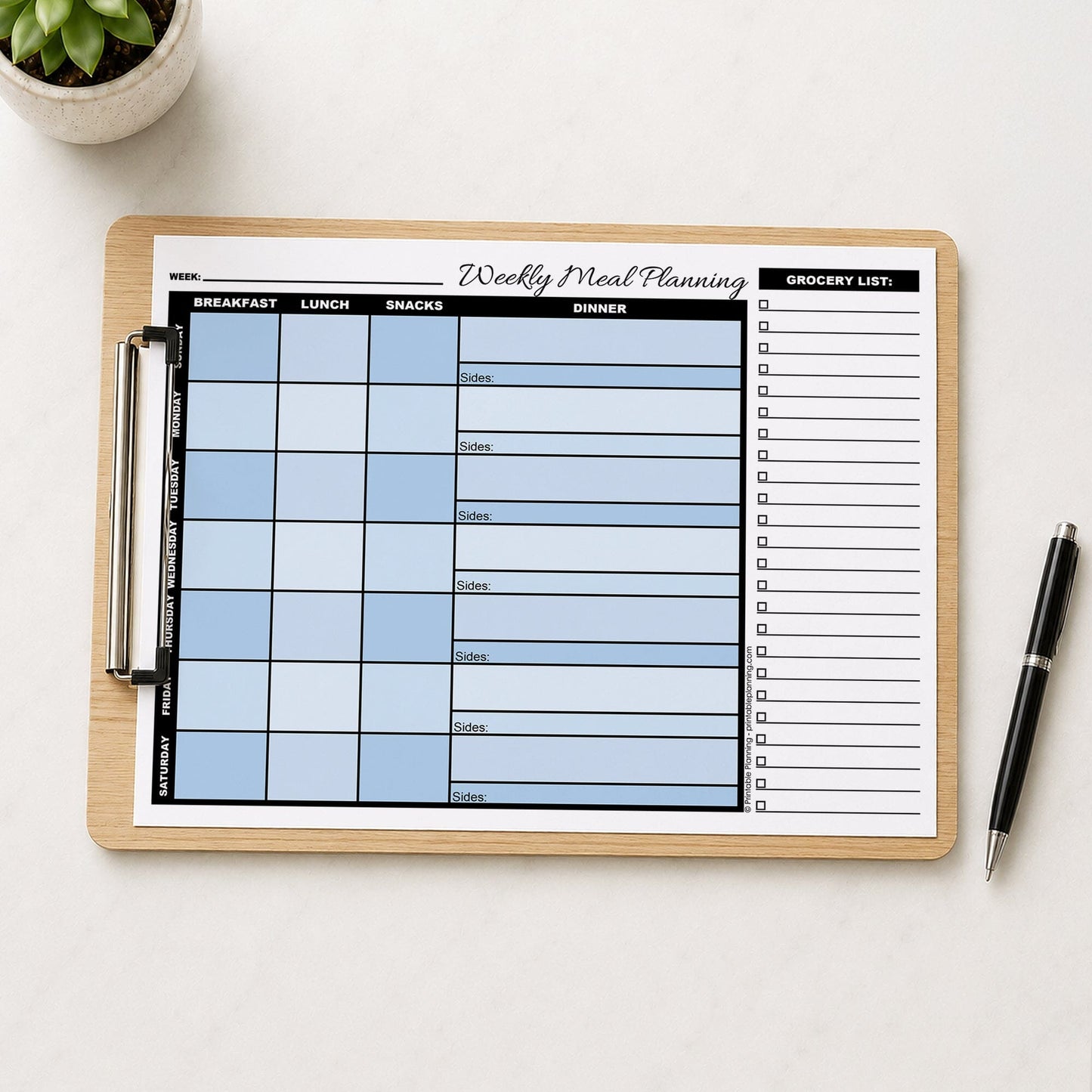 A printed Blue Weekly Meal Planner with Grocery List (printable page) shown on a wooden clipboard on a table with a plant and a pen.