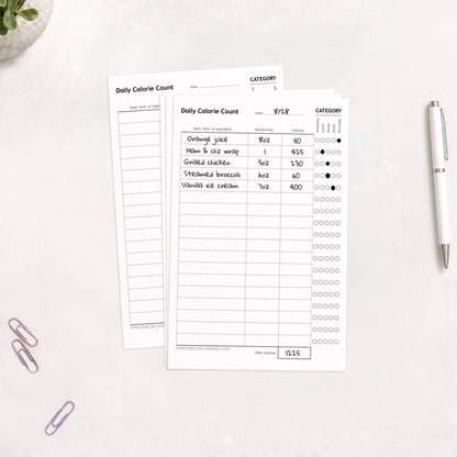 Printed pages of the printable Daily Calorie Tracker & Food Log (Half-Page), shown with example writing, on a table with a plant, paperclips, and a pen.