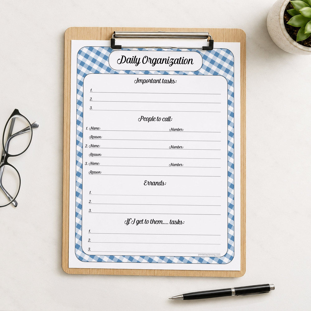 The printed Blue Gingham Daily Planner with Priorities, Calls, Errands & Tasks (printable) page shown on a wooden clipboard on a table with a plant, glasses, and a pen.