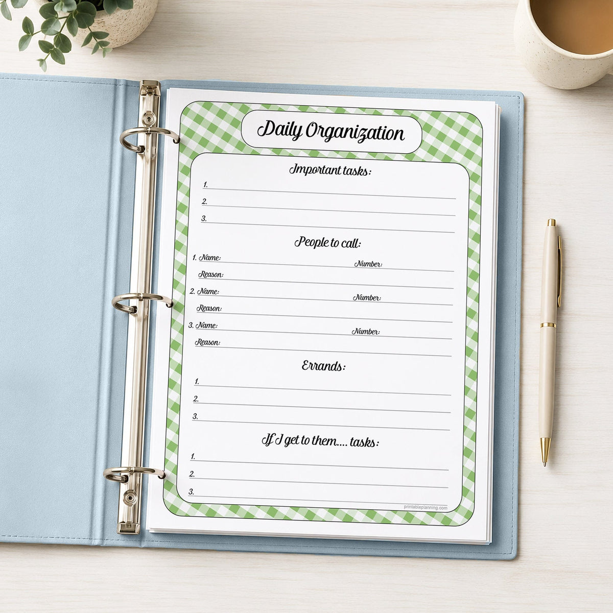 The printed green gingham Daily Organization Planner with Priorities, Calls & Errands (printable) page shown in a light blue binder on a desk with a plant, coffee, and a pen.