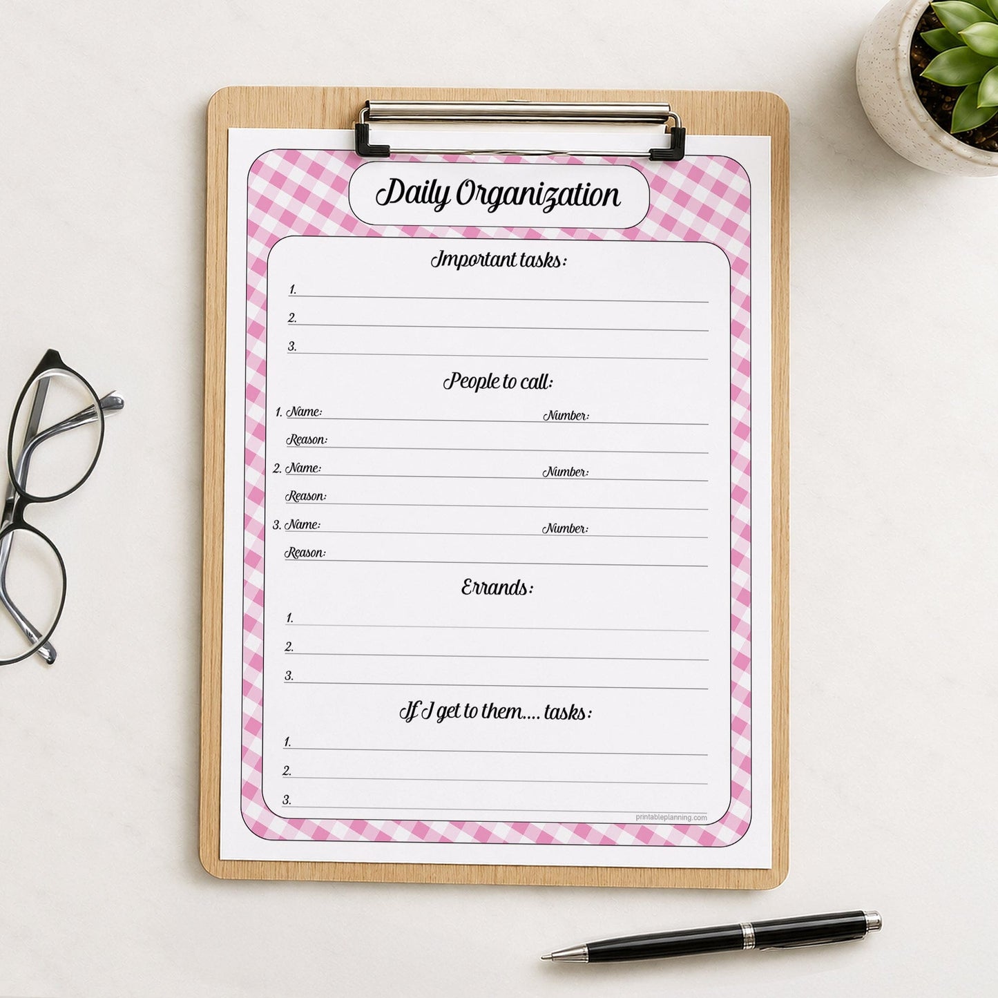 The printed pink gingham Daily Planner with Priorities, Calls & Errands (printable) page shown on a wooden clipboard on a table with a plant, glasses, and a pen.