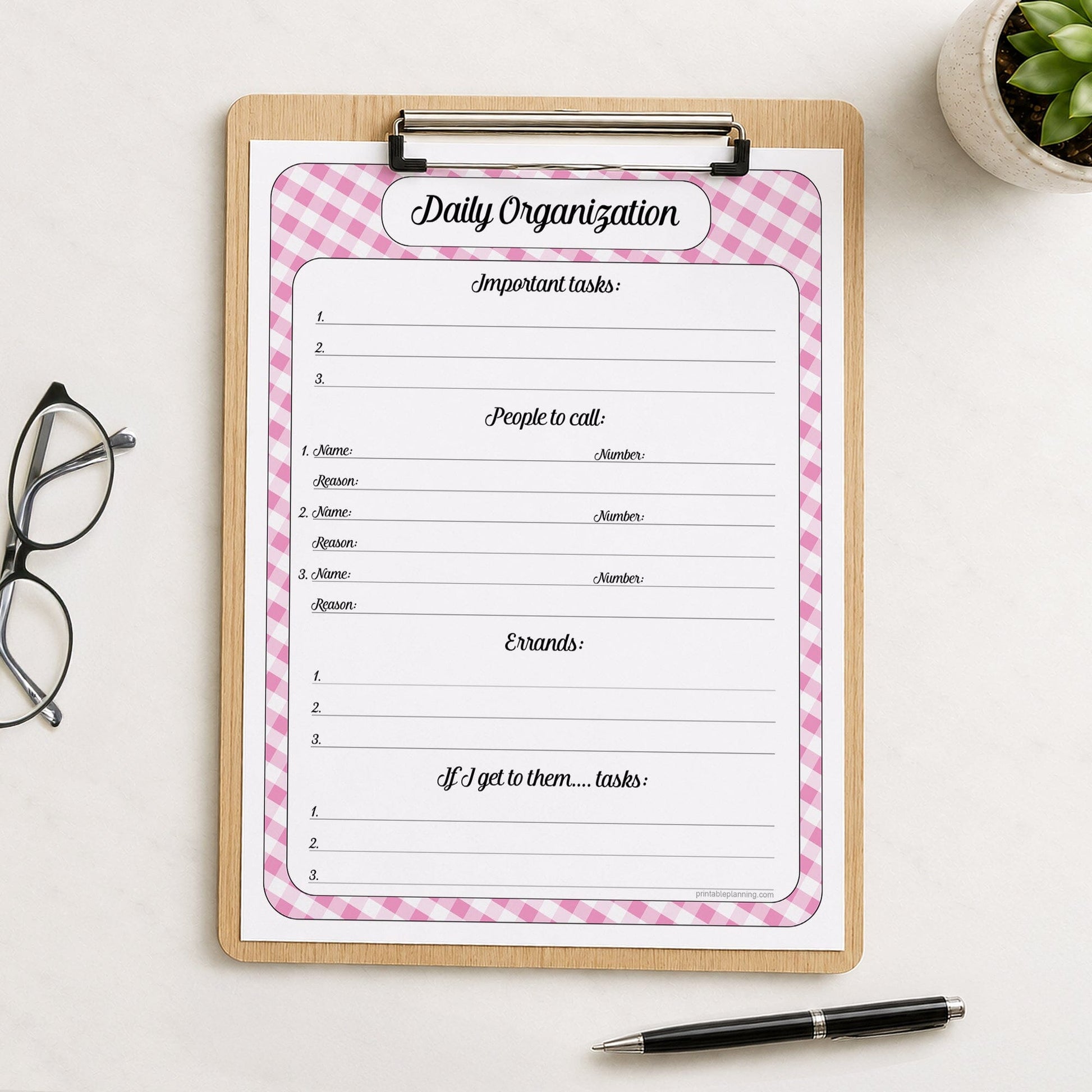 The printed pink gingham Daily Planner with Priorities, Calls & Errands (printable) page shown on a wooden clipboard on a table with a plant, glasses, and a pen.
