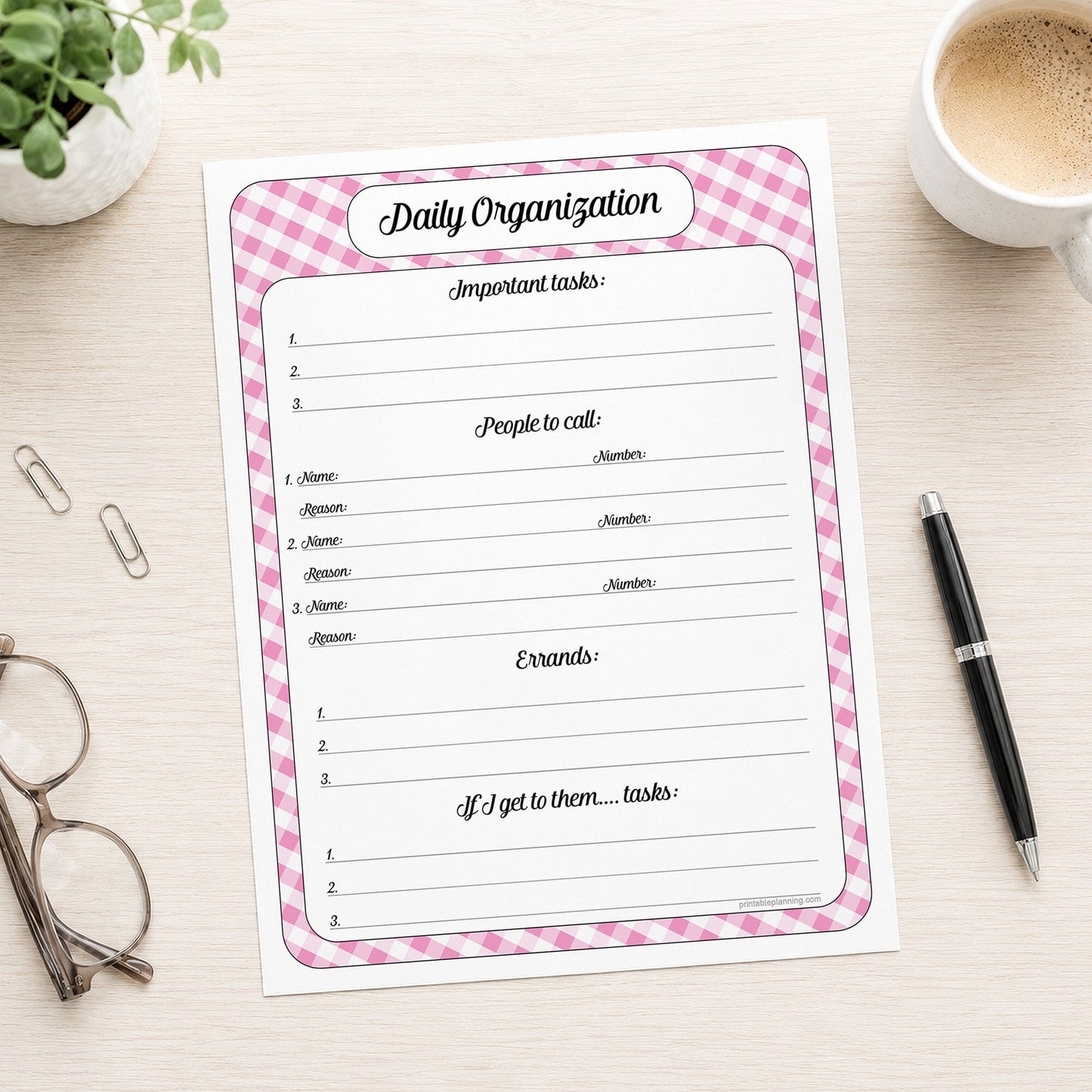 The printed pink gingham Daily Planner with Priorities, Calls & Errands (printable) page shown on a desk with a plant, glasses, paperclips, coffee, and a pen.