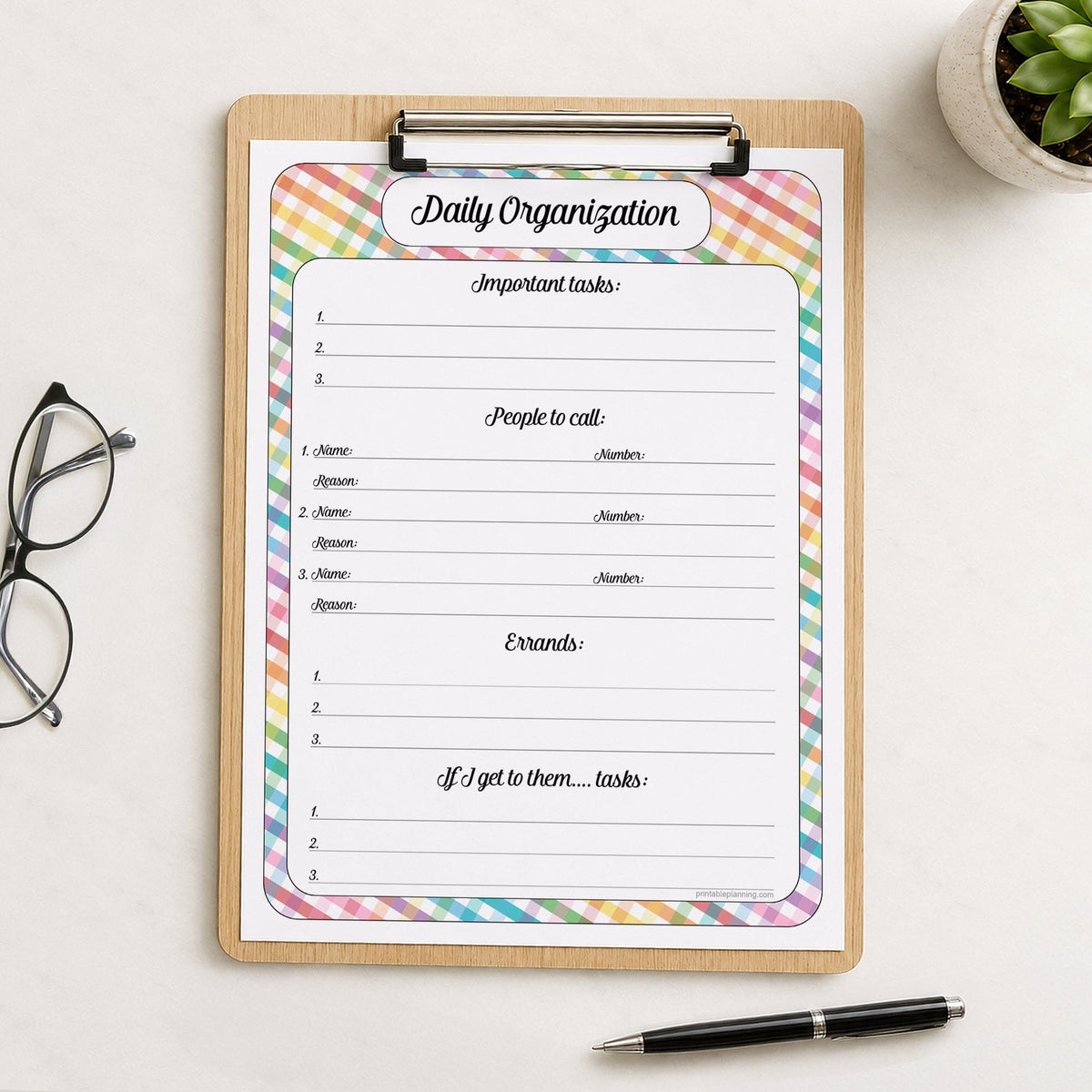A printed rainbow gingham Daily Planner for Priorities, Calls, Errands & Tasks (printable) page shown on a wooden clipboard on a table with a plant, glasses, and a pen.