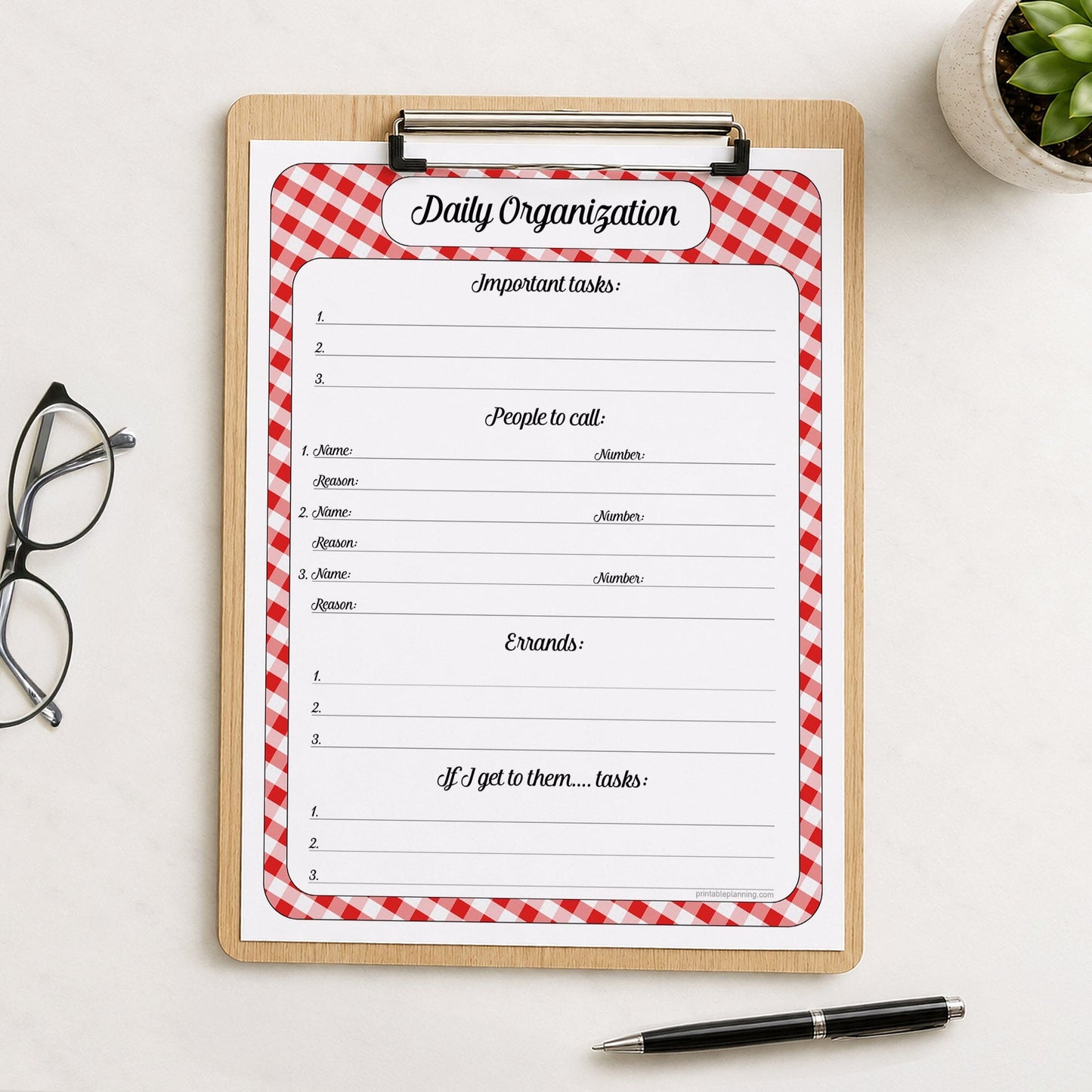 A printed red gingham Daily Priority Planner with Calls, Errands & Tasks (printable) page shown on a wooden clipboard on a table with a plant, glasses, and a pen.