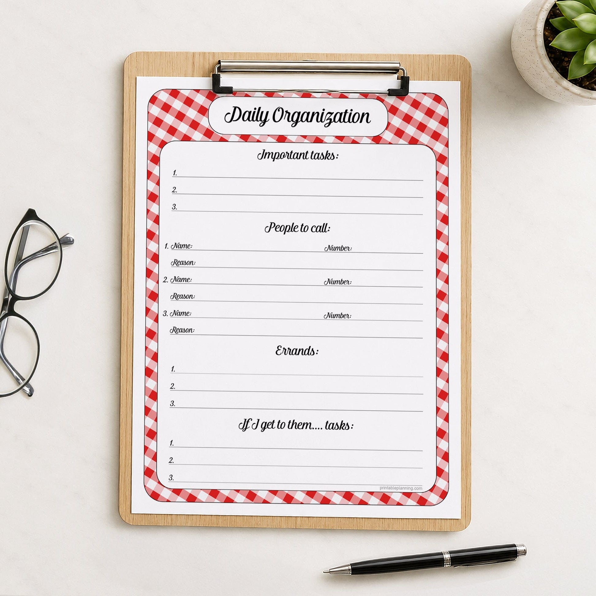 A printed red gingham Daily Priority Planner with Calls, Errands & Tasks (printable) page shown on a wooden clipboard on a table with a plant, glasses, and a pen.