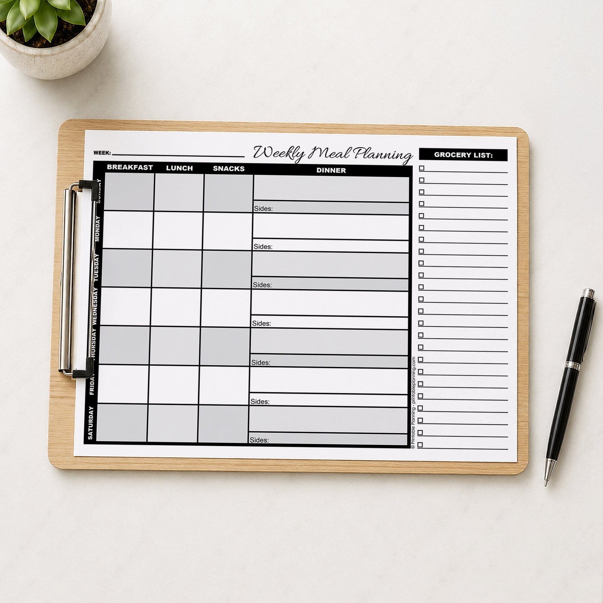A printed Grayscale Weekly Meal Planner with Grocery List (printable page) shown on a wooden clipboard on a table with a plant and a pen.
