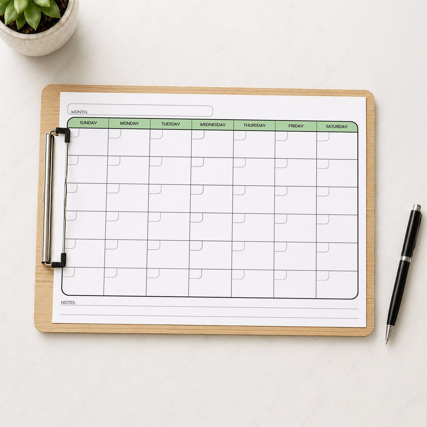 The printed Undated Monthly Calendar - Green Full-Page Planner (printable) page shown on a wooden clipboard on a table with a plant and a pen.