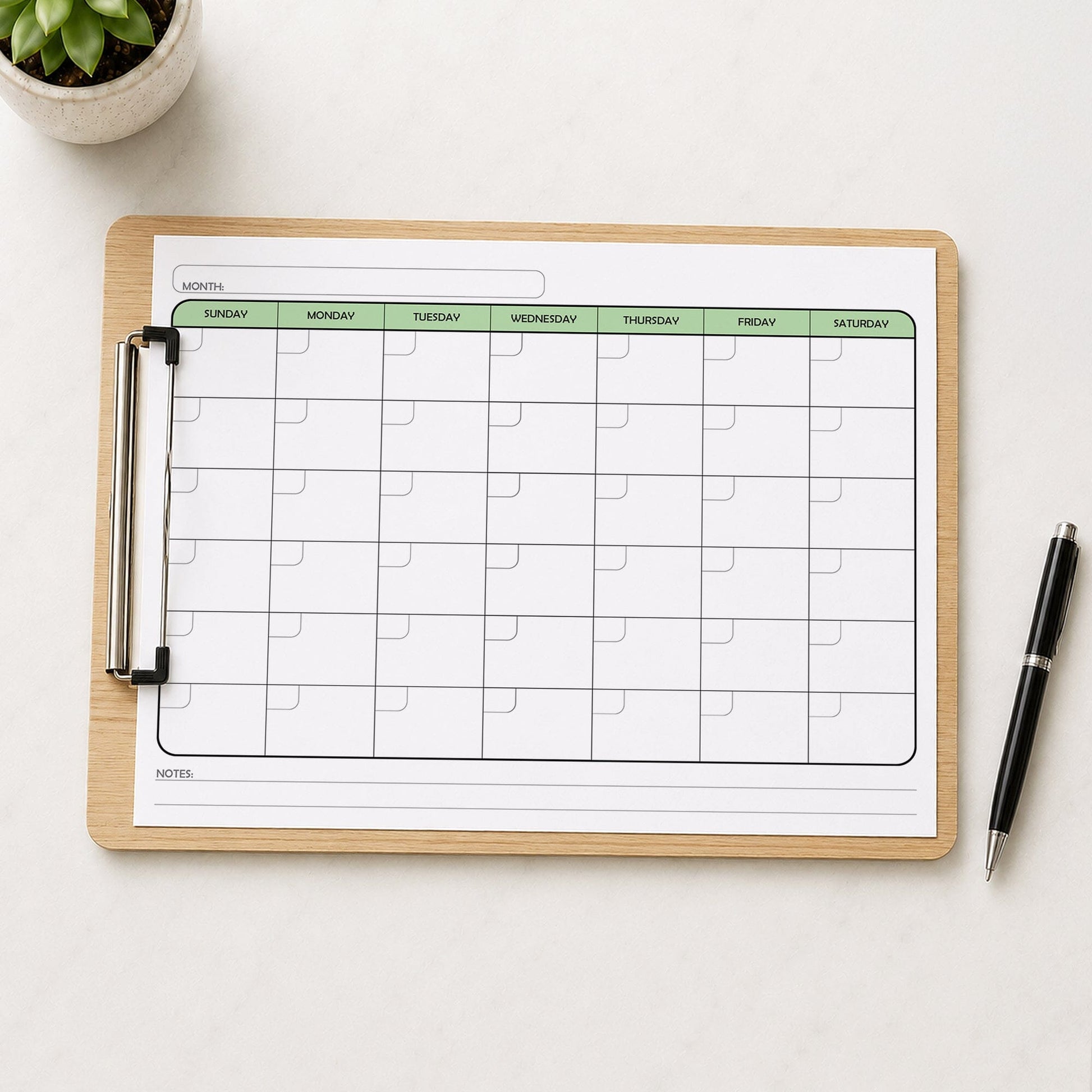 The printed Undated Monthly Calendar - Green Full-Page Planner (printable) page shown on a wooden clipboard on a table with a plant and a pen.