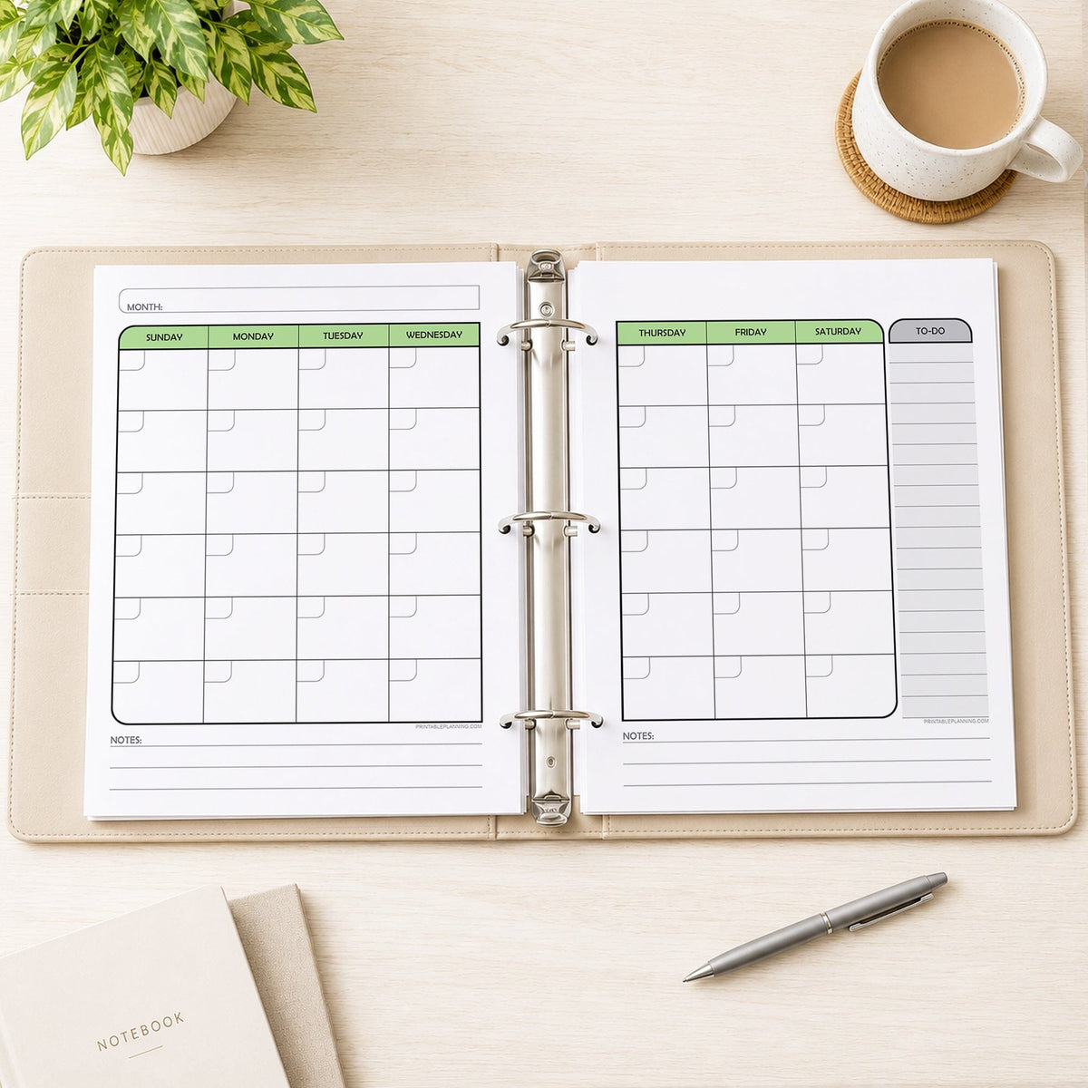 The printed Green Undated Monthly Planner (printable) Pages shown in an open beige binder as facing pages, on a desk with a plant, coffee, notebooks, and a pen.