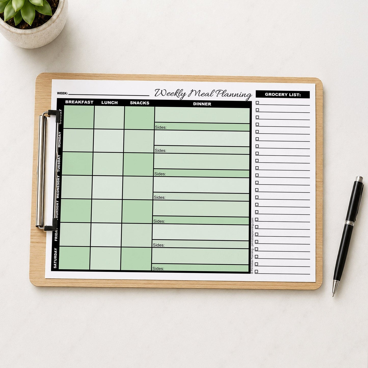 A printed Green Weekly Meal Planner with Grocery List (printable page) shown on wooden clipboard on a table with a plant and a pen.