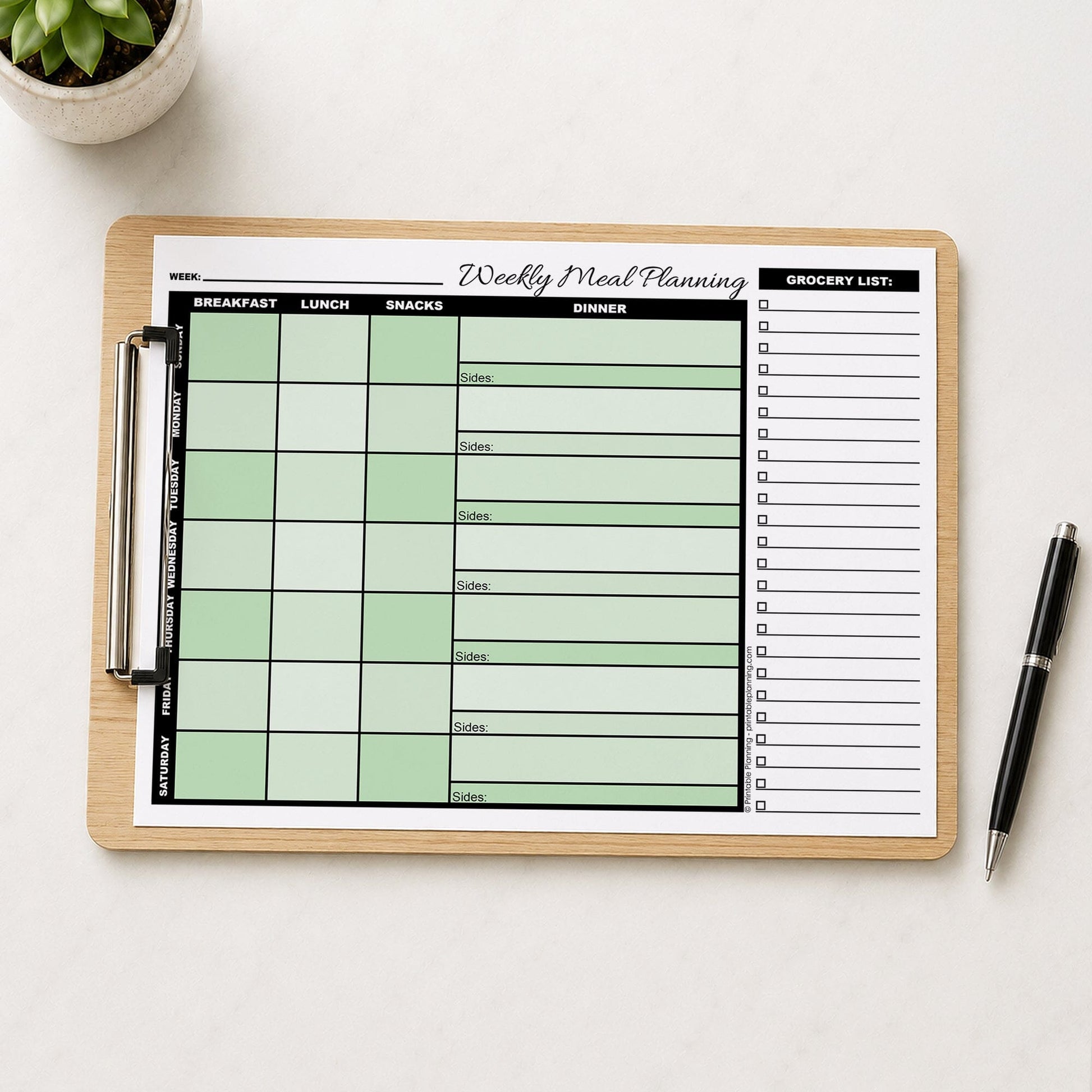 A printed Green Weekly Meal Planner with Grocery List (printable page) shown on wooden clipboard on a table with a plant and a pen.