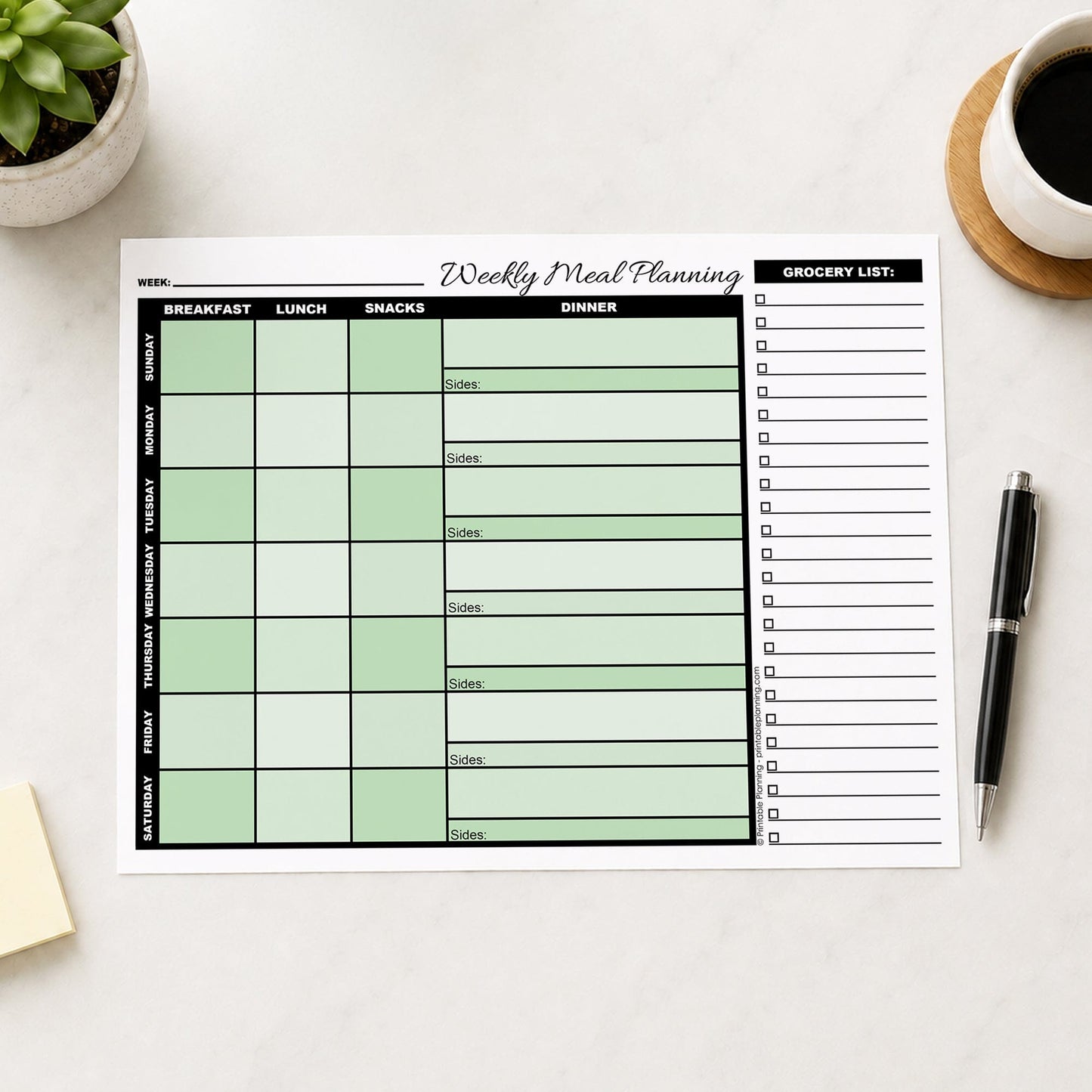 A printed Green Weekly Meal Planner with Grocery List (printable page) shown on a desk with a plant, sticky notes, coffee, and a pen.