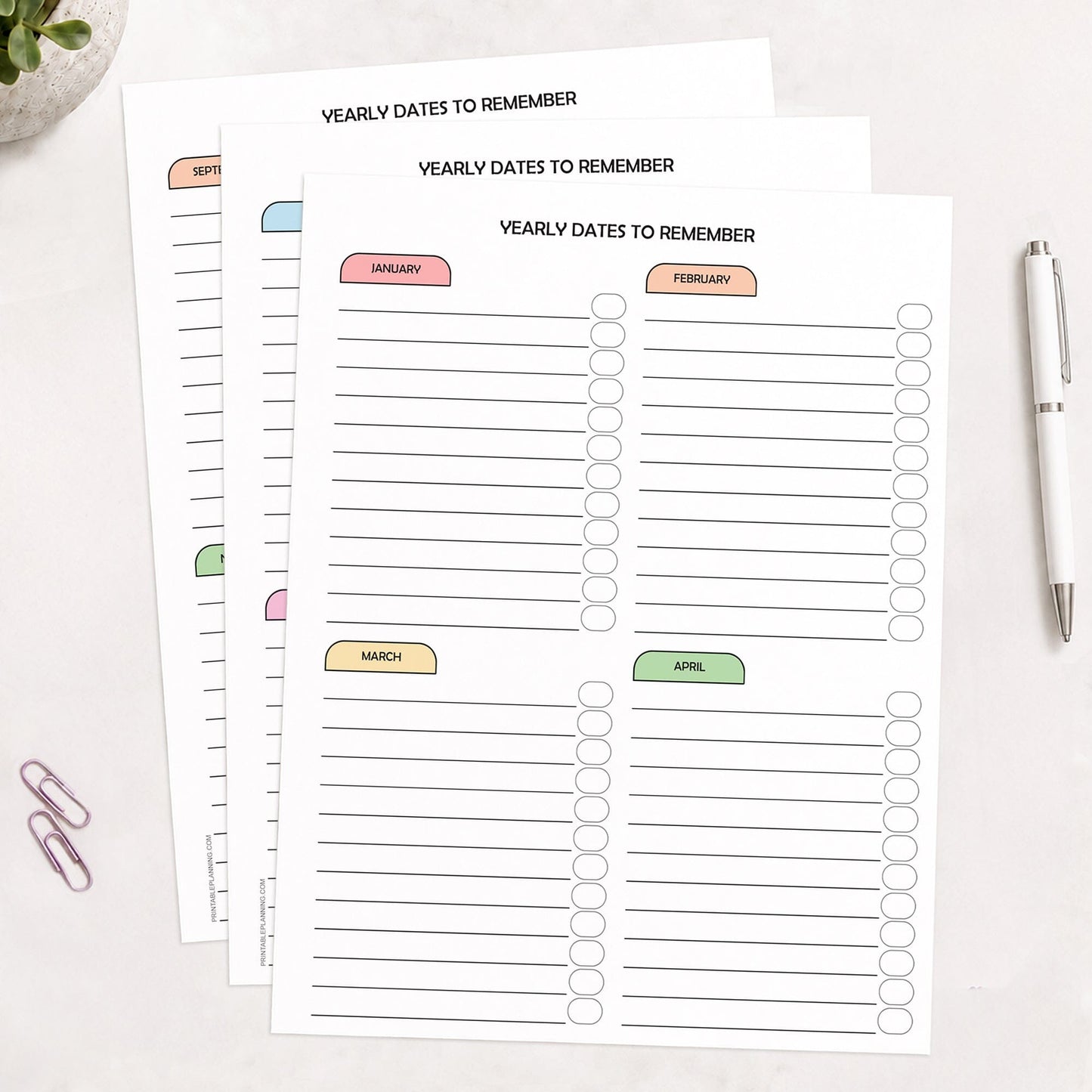 Three printed (printable) planner pages titled 'YEARLY DATES TO REMEMBER' arranged on a light surface, with monthly sections labeled January through December across all pages, in pastel colors in a rainbow progression. A white pen and two paperclips are placed nearby as styled props.
