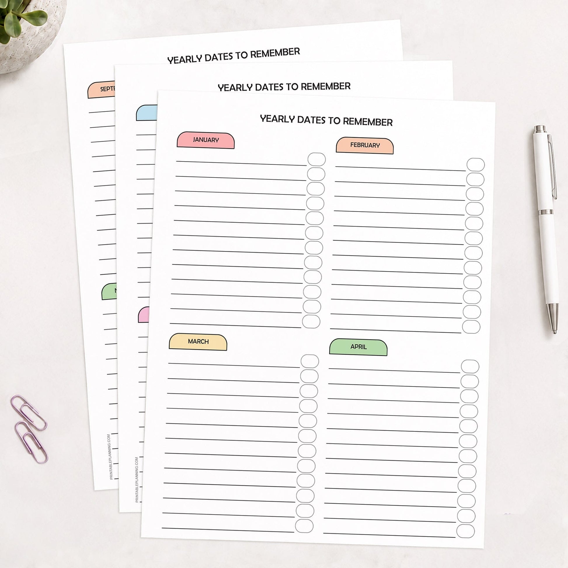 Three printed (printable) planner pages titled 'YEARLY DATES TO REMEMBER' arranged on a light surface, with monthly sections labeled January through December across all pages, in pastel colors in a rainbow progression. A white pen and two paperclips are placed nearby as styled props.