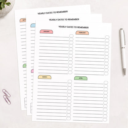 Three printed (printable) planner pages titled 'YEARLY DATES TO REMEMBER' arranged on a light surface, with monthly sections labeled January through December across all pages, in pastel colors in a rainbow progression. A white pen and two paperclips are placed nearby as styled props.