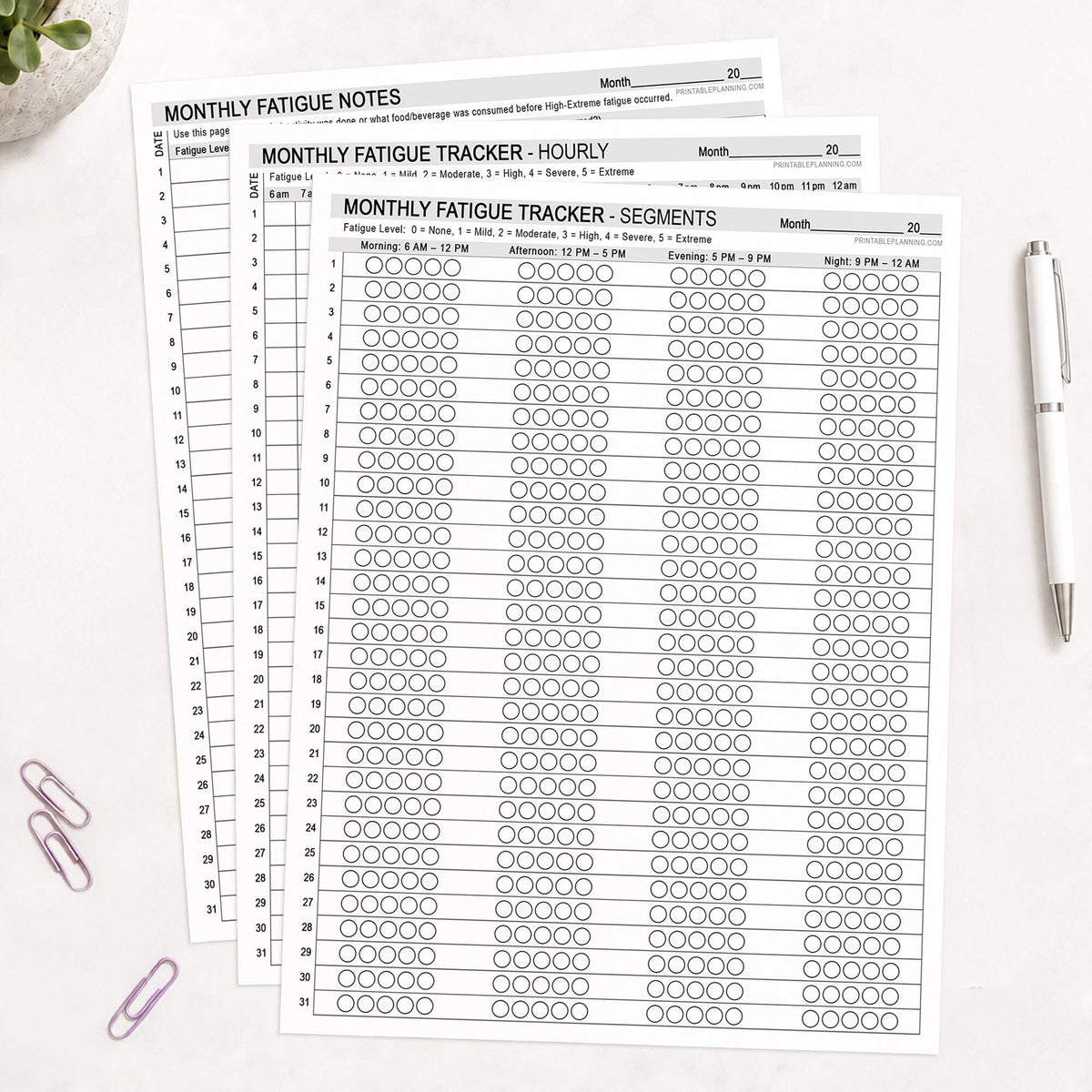 Three printed monthly fatigue tracker sheets arranged in a fan shape on a white surface, with a white pen, pink paperclips, and a small plant visible around them.