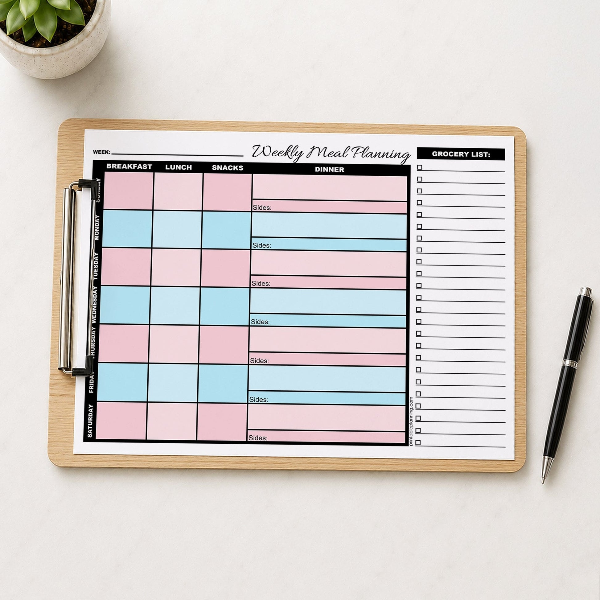 A printed Pink Blue Weekly Meal Planner with Grocery List (printable page) shown on a wooden clipboard on a table with a plant and a pen.