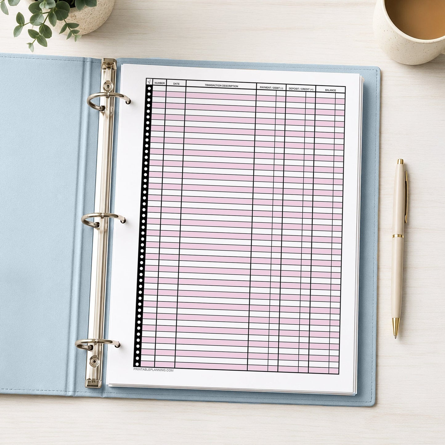 Pink Printable Check Register - Full-Page Transaction Tracker shown in a light blue binder on a desk with a plant, coffee, and a pen.