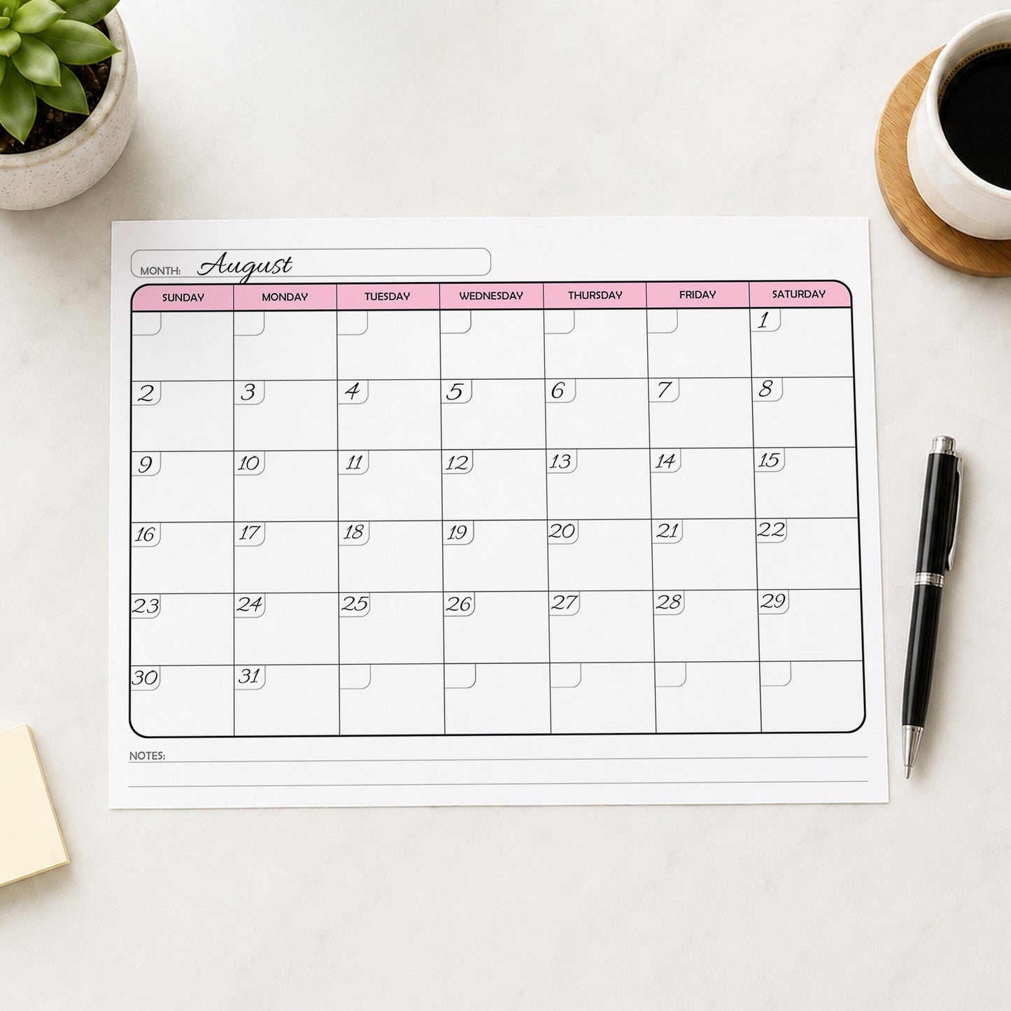 A printed Undated Monthly Calendar - Pink Full-Page Planner (printable) page shown with example writing on a desk with a plant, coffee, sticky notes, and a pen.