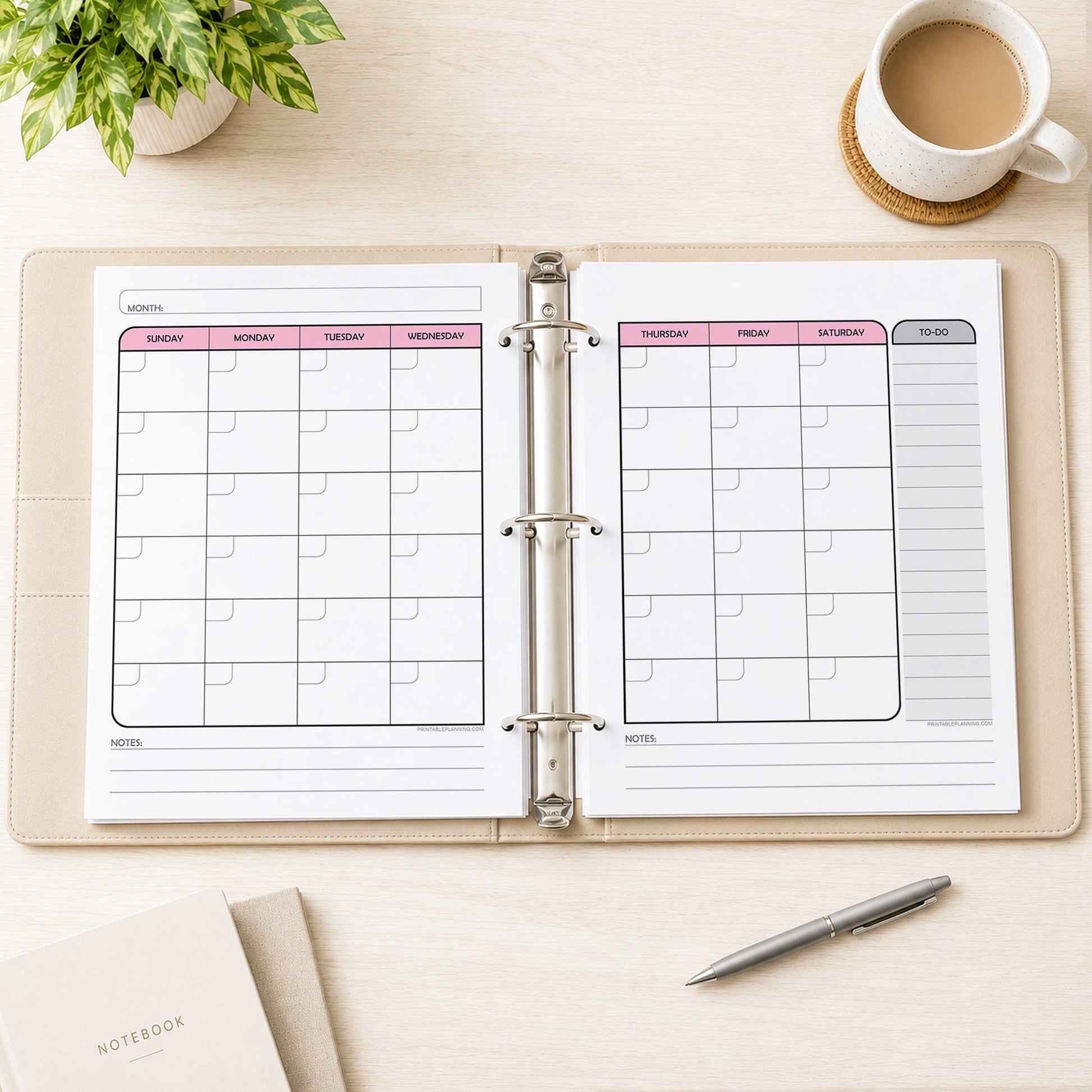 The printed Pink Undated Monthly Planner (printable) Pages shown in an open beige binder showing facing pages, on a desk with a plant, coffee, notebooks, and a pen.