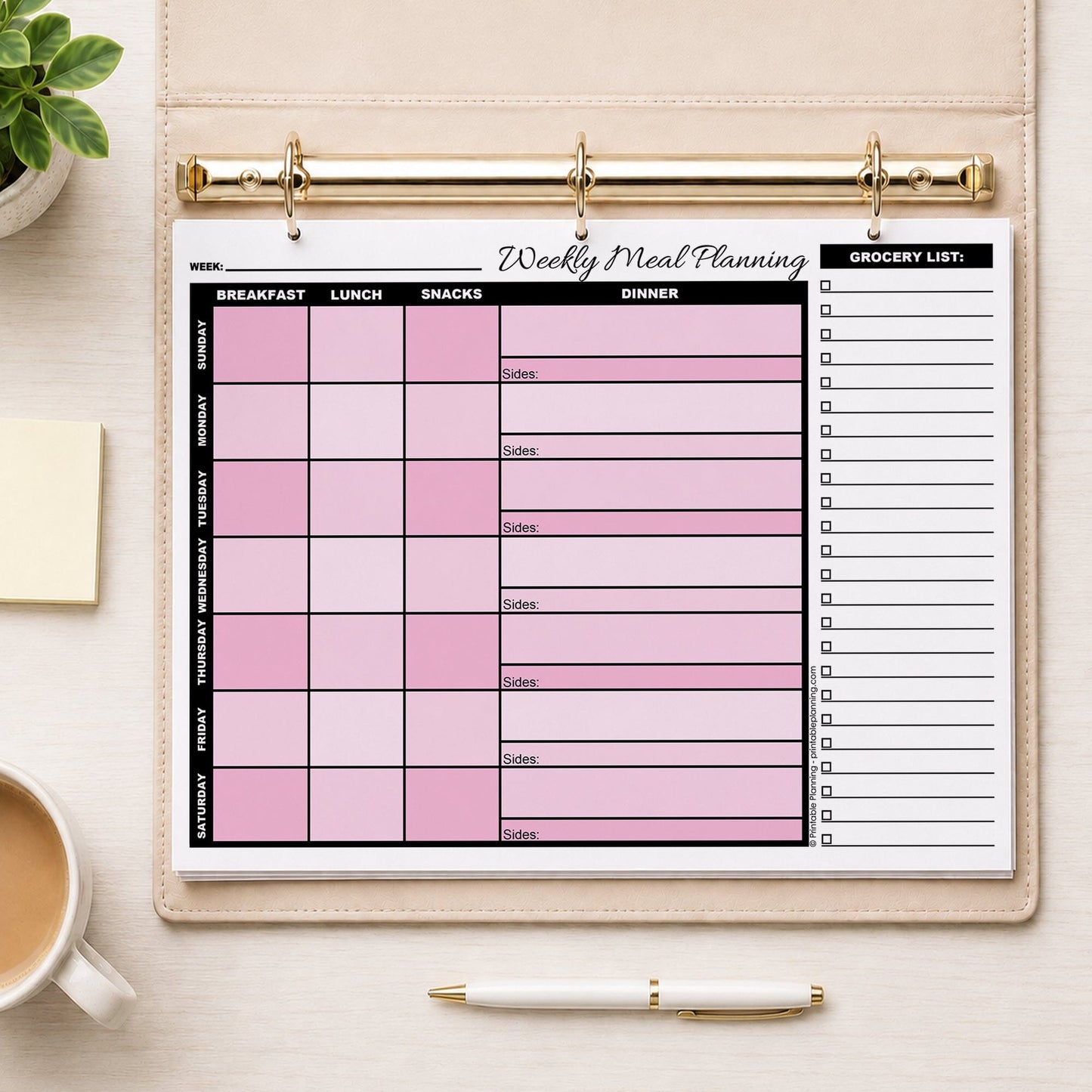 A printed Pink Weekly Meal Planner with Grocery List (printable page) shown in a beige binder on a table with a plant, sticky notes, coffee, and a pen.