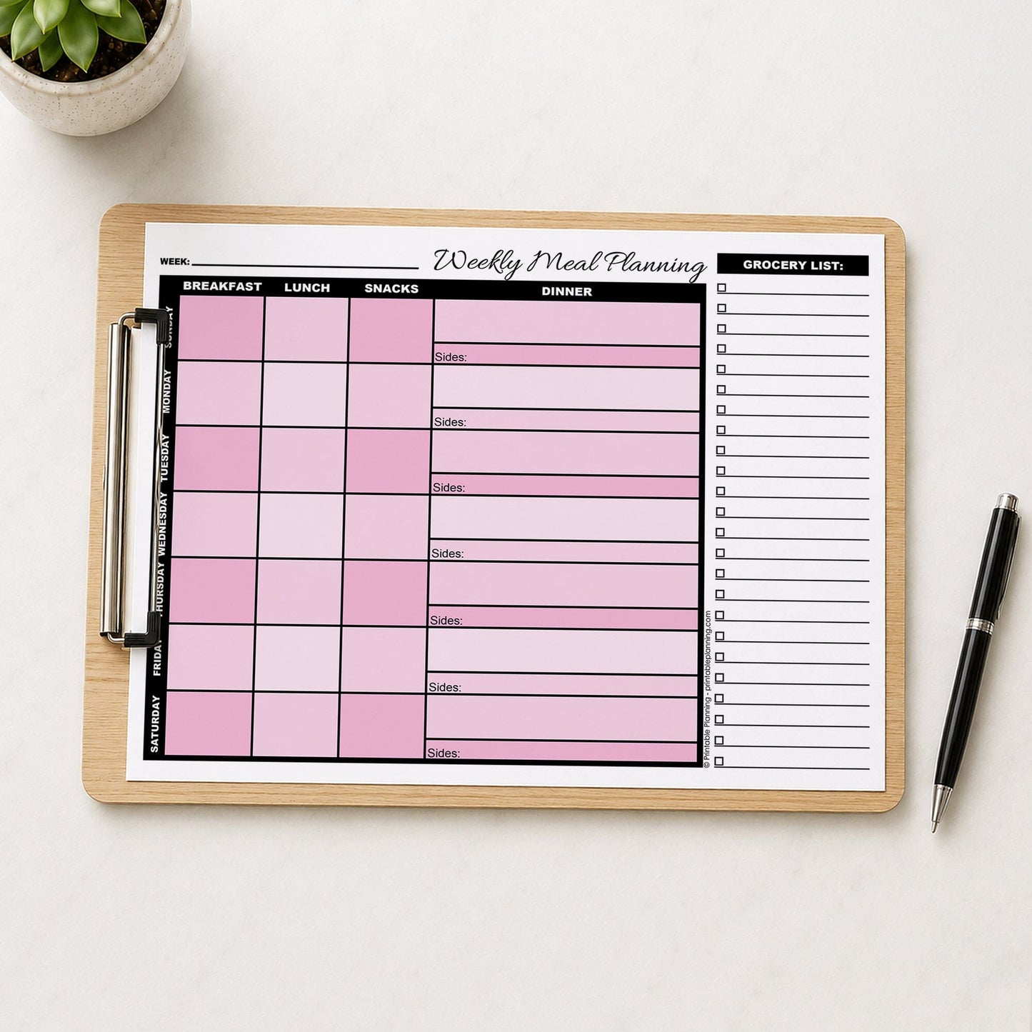 A printed Pink Weekly Meal Planner with Grocery List (printable page) shown on a wooden clipboard on a table with a plant, and a pen.