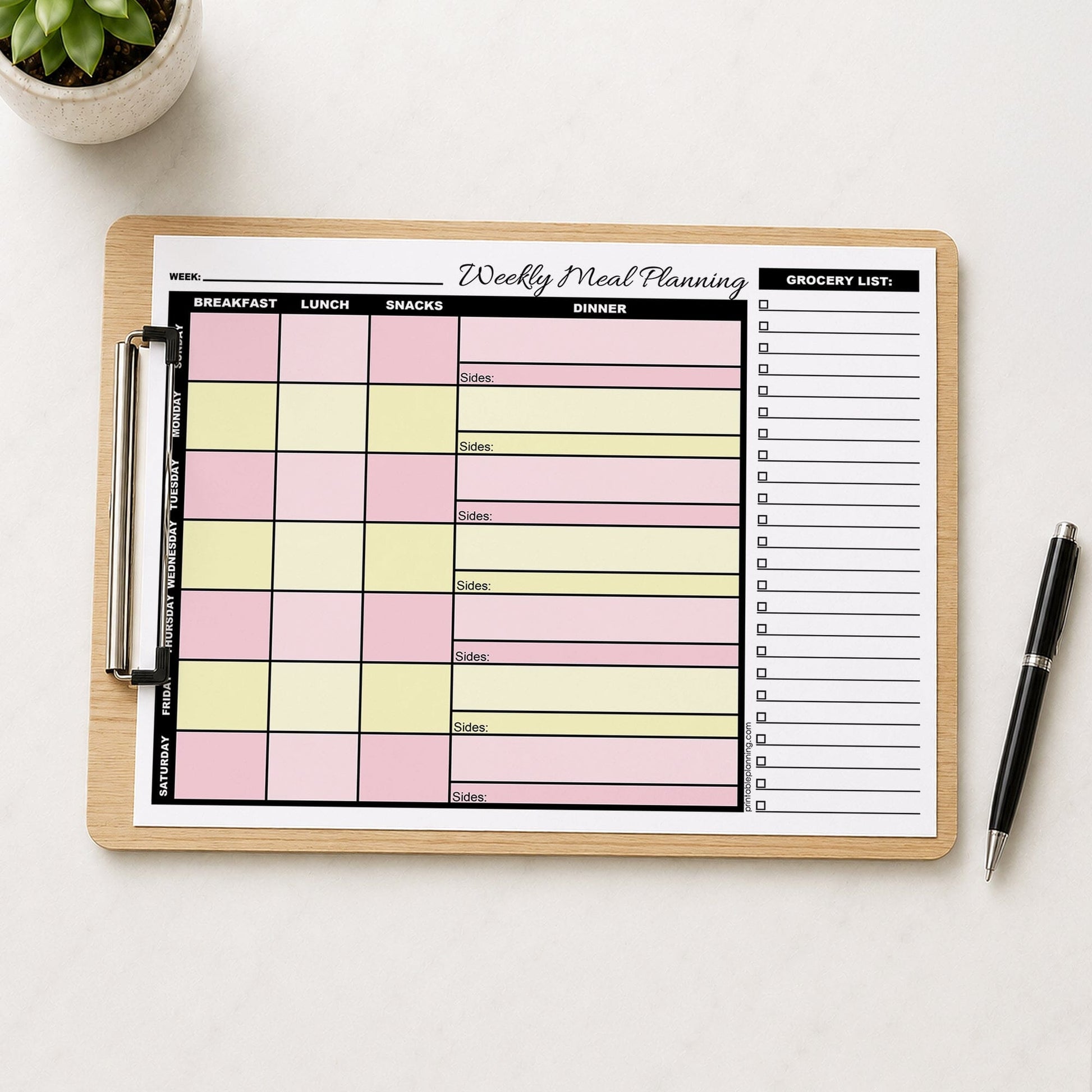 A printed Pink Yellow Weekly Meal Planner with Grocery List (printable) page displayed on a wooden clipboard, styled on a light table surface with a small potted succulent in the top-left corner and a black pen on the right.