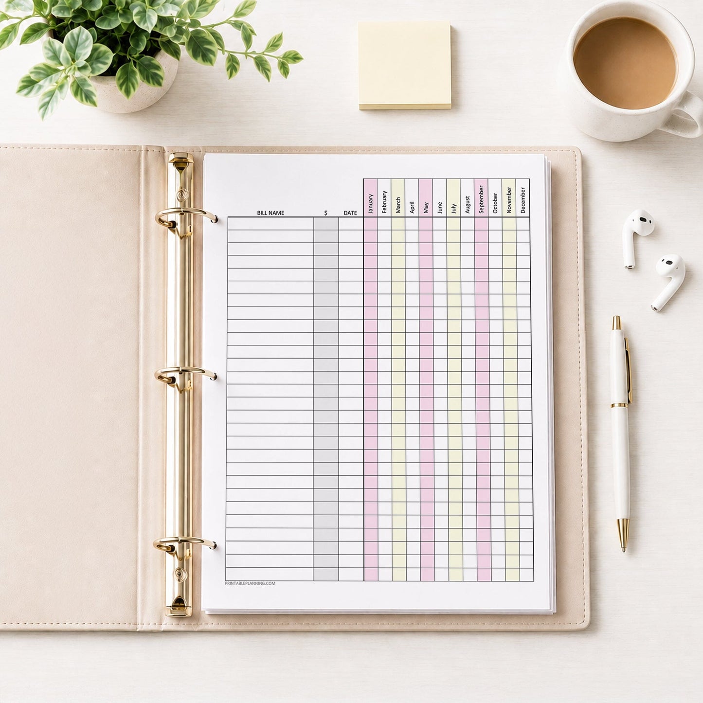 Pink Yellow Yearly Bill Payment Tracker with Amount Column (printable page) shown in a beige binder with a plant, earbuds, notes, coffee, and a pen.