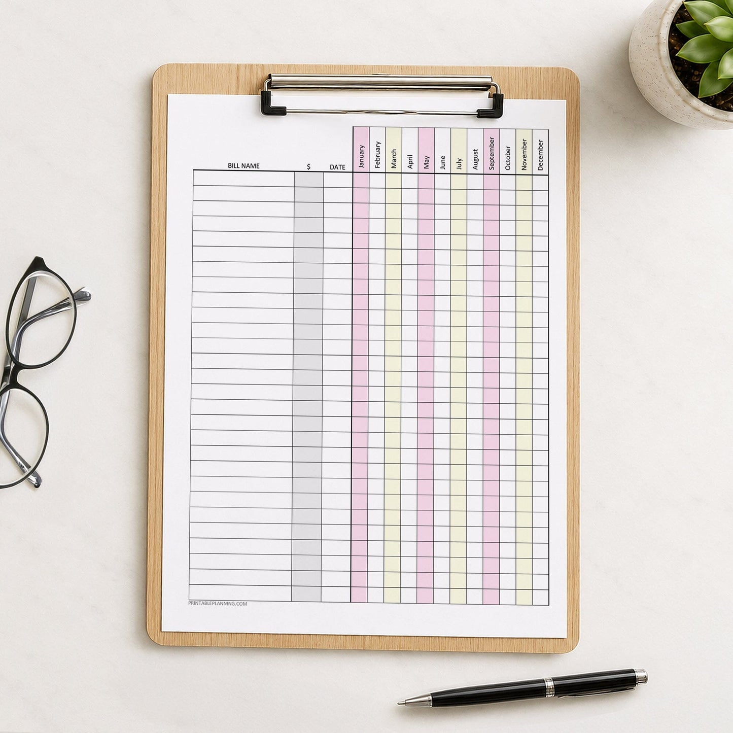 Pink Yellow Yearly Bill Payment Tracker with Amount Column (printable page) shown on a wooden clipboard on a desk with a plant, glasses, and a pen.