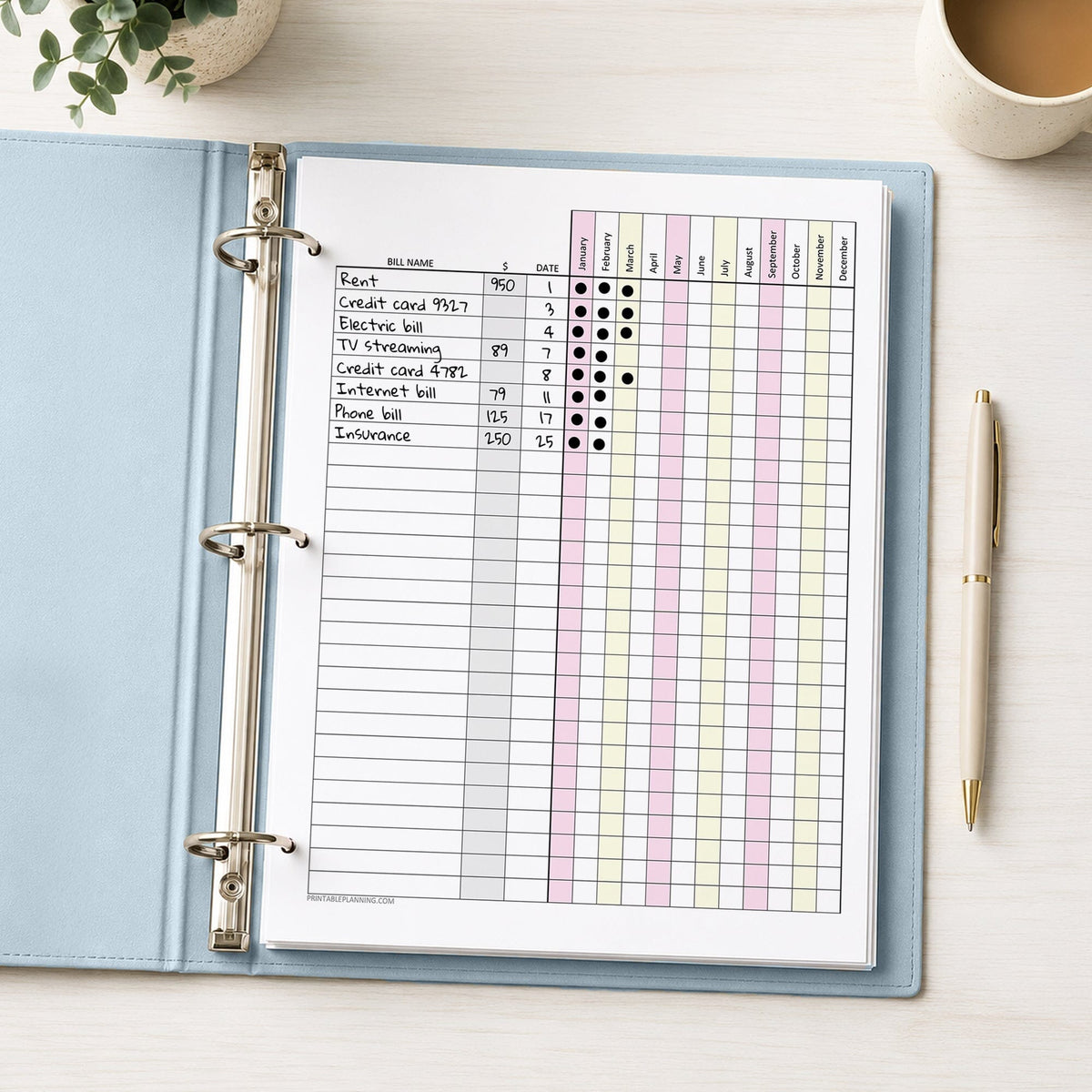 A printed Pink Yellow Yearly Bill Payment Tracker with Amount Column (printable) page, shown with example writing, in a light blue binder on a desk with a plant, coffee, and a pen.