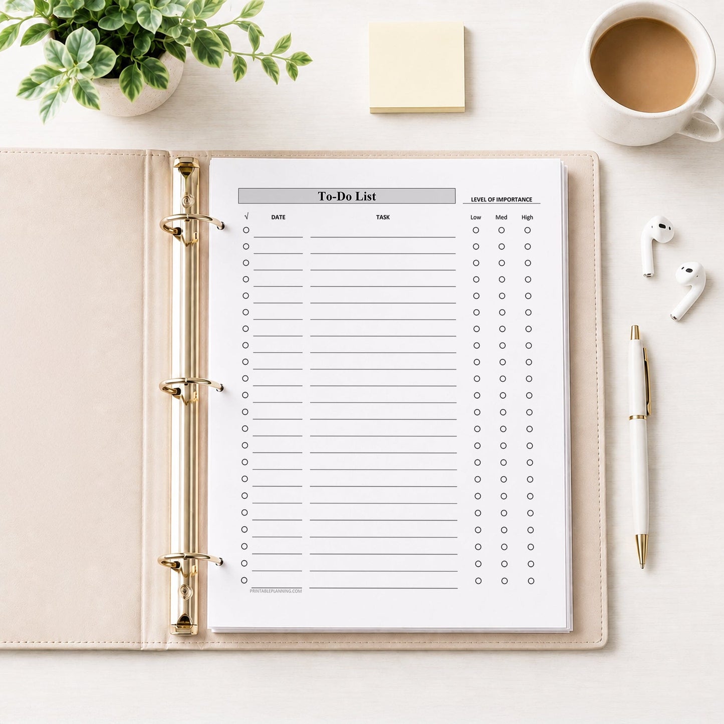 A flat-lay view of a printed Priority To-Do List with Low Medium High Importance - Printable PDF page inside a beige leather ring binder, surrounded by lifestyle props including a cup of coffee, white earbuds, a white and pen, and a small potted succulent on a white desk surface.