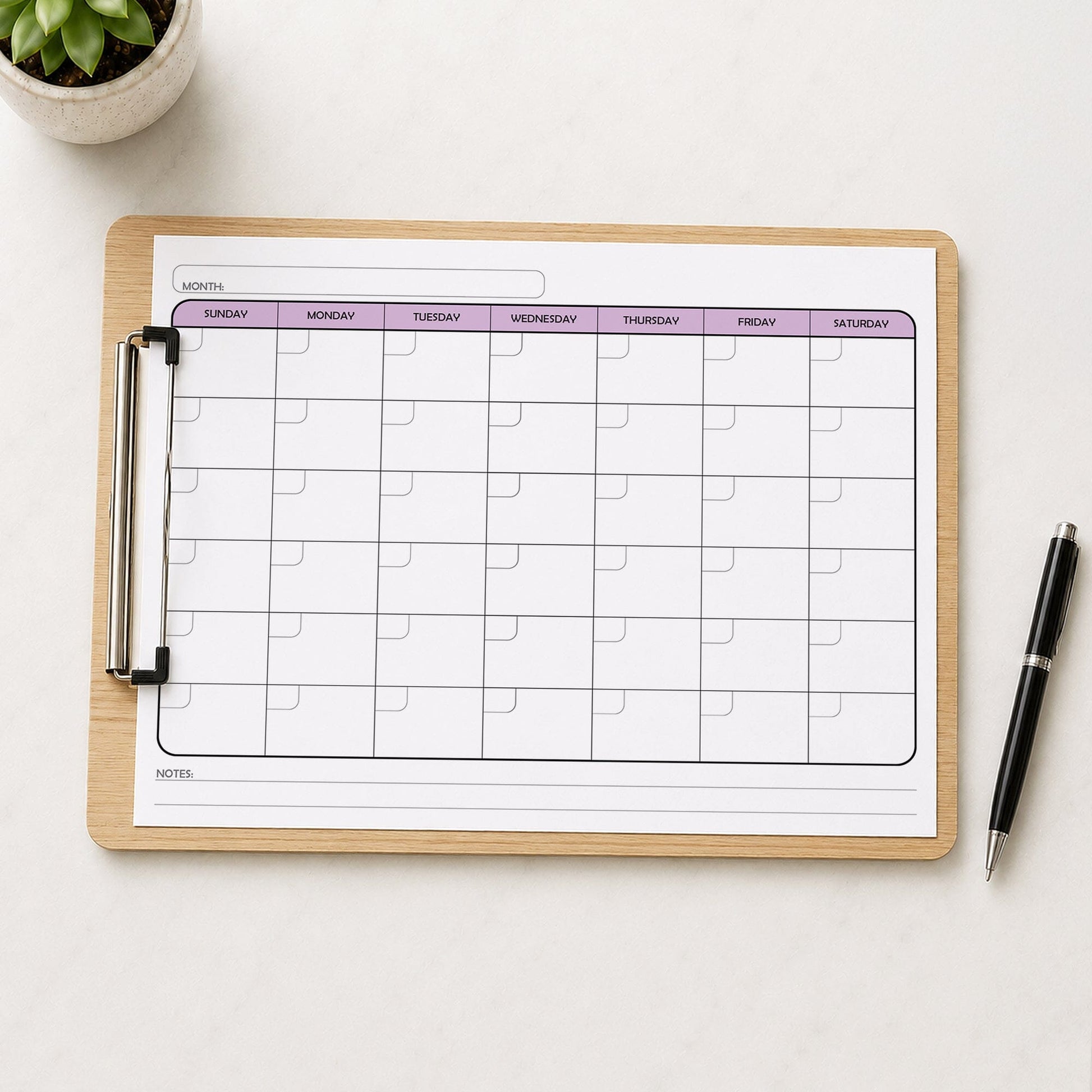 The printed Undated Monthly Calendar - Purple Full-Page Planner (printable) page shown on a wooden clipboard on a table with a plant and a pen.