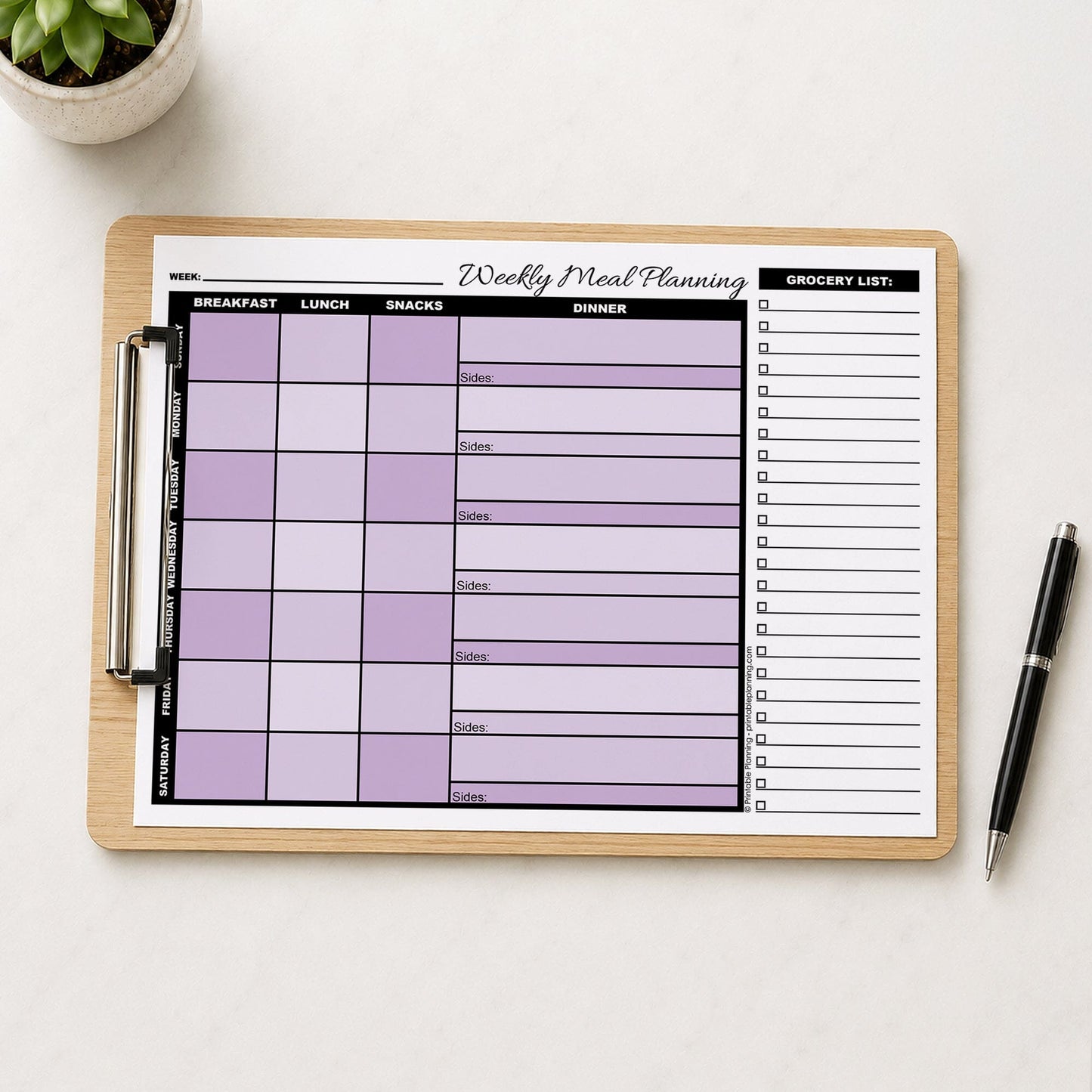 A printed Purple Weekly Meal Planner with Grocery List (printable page) shown shown on a wooden clipboard on a table with a plant and a pen.