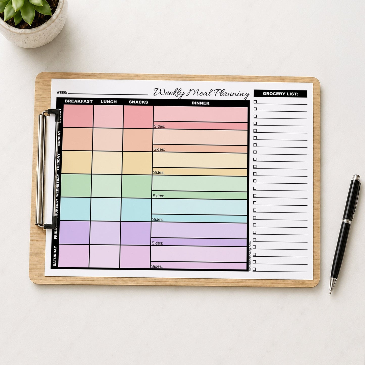 A Rainbow Weekly Meal Planner with Grocery List (printable page) shown on a wooden clipboard on a table with a plant and a pen.