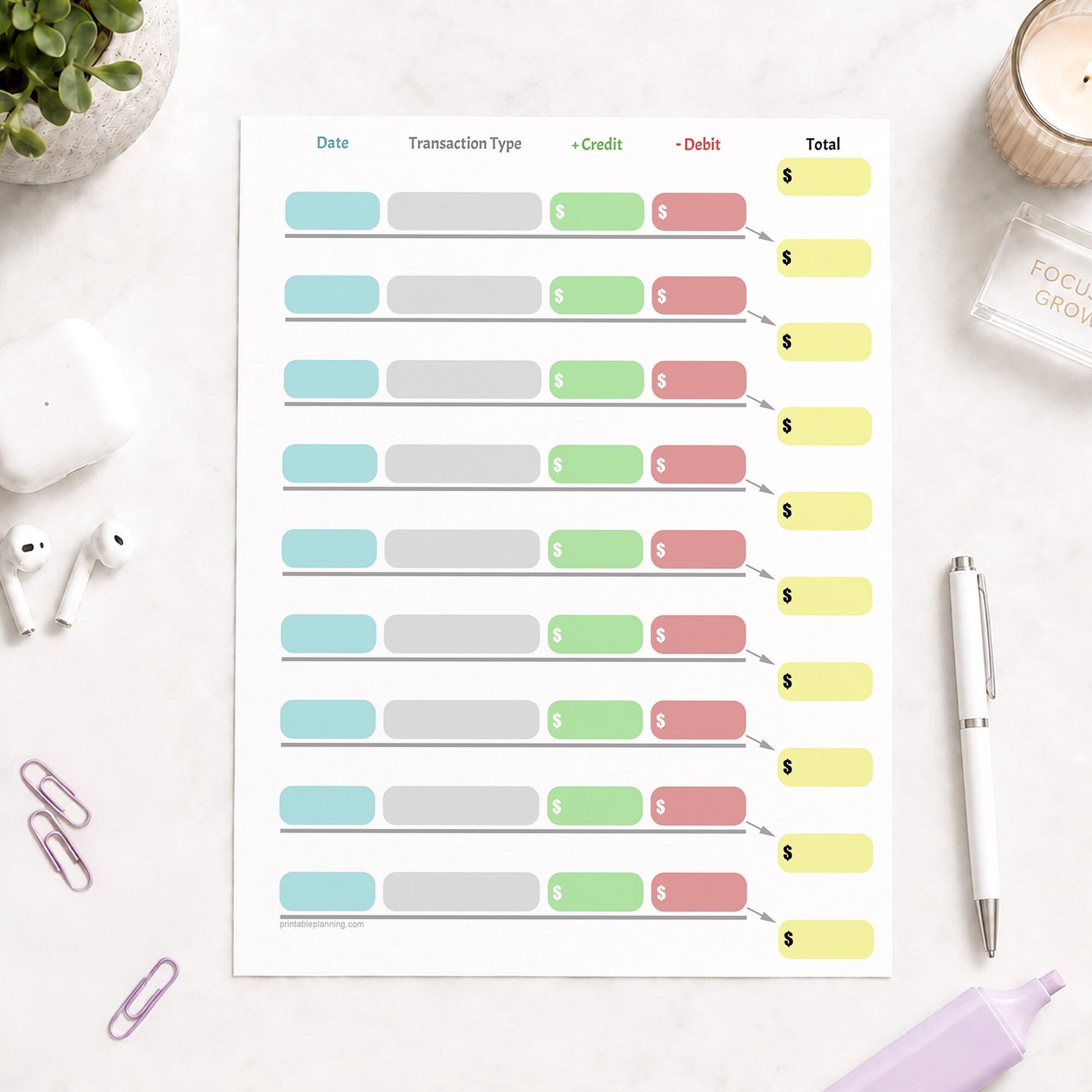 Simple Color-Coded Financial Transaction Register (printable page) shown on a light desk with earbuds, paperclips, highlighter, plant, and a pen.