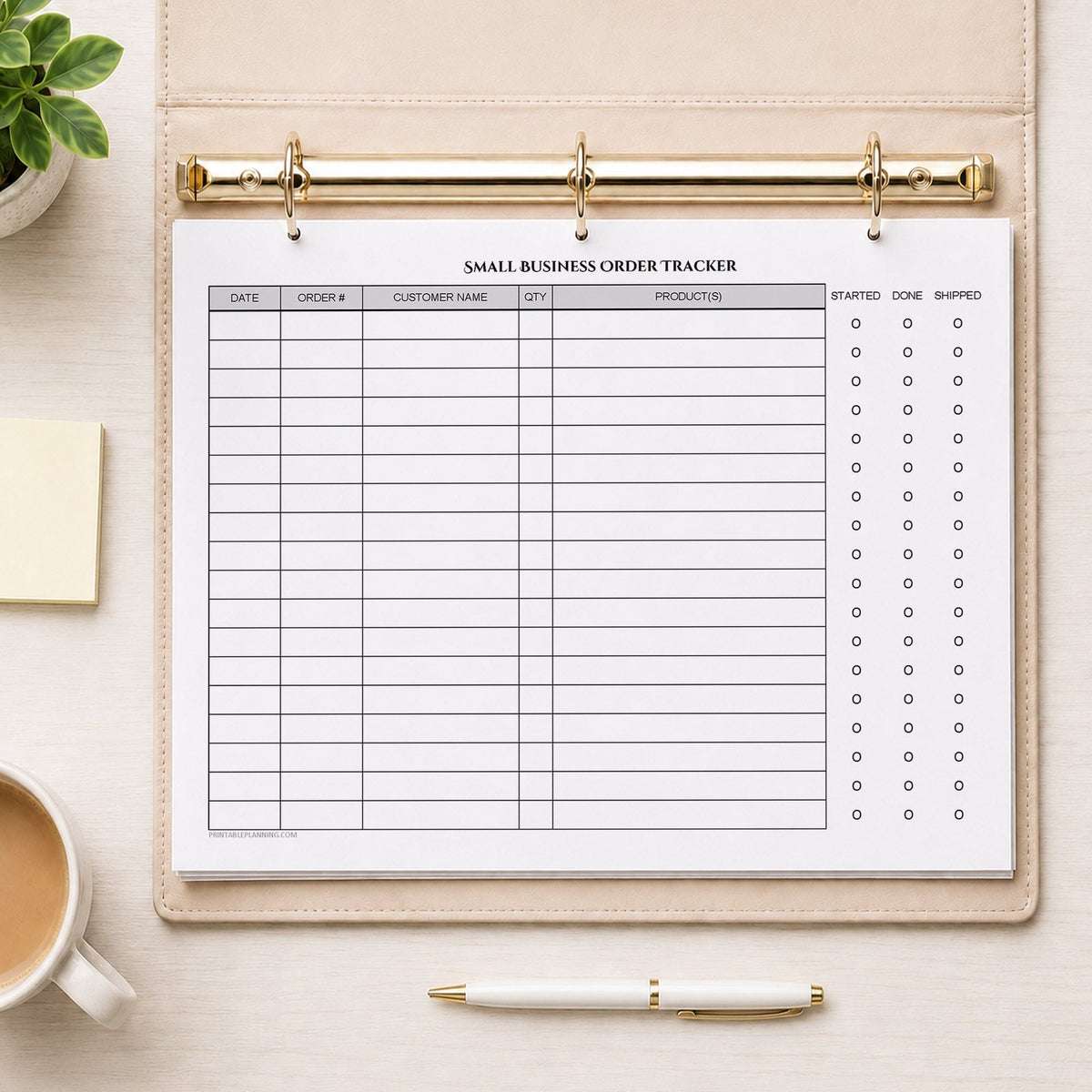 A printed Small Business Order Tracker with Order Status Checklist (printable) page shown in a beige binder on a desk with a plant, sticky notes, coffee, and a pen.