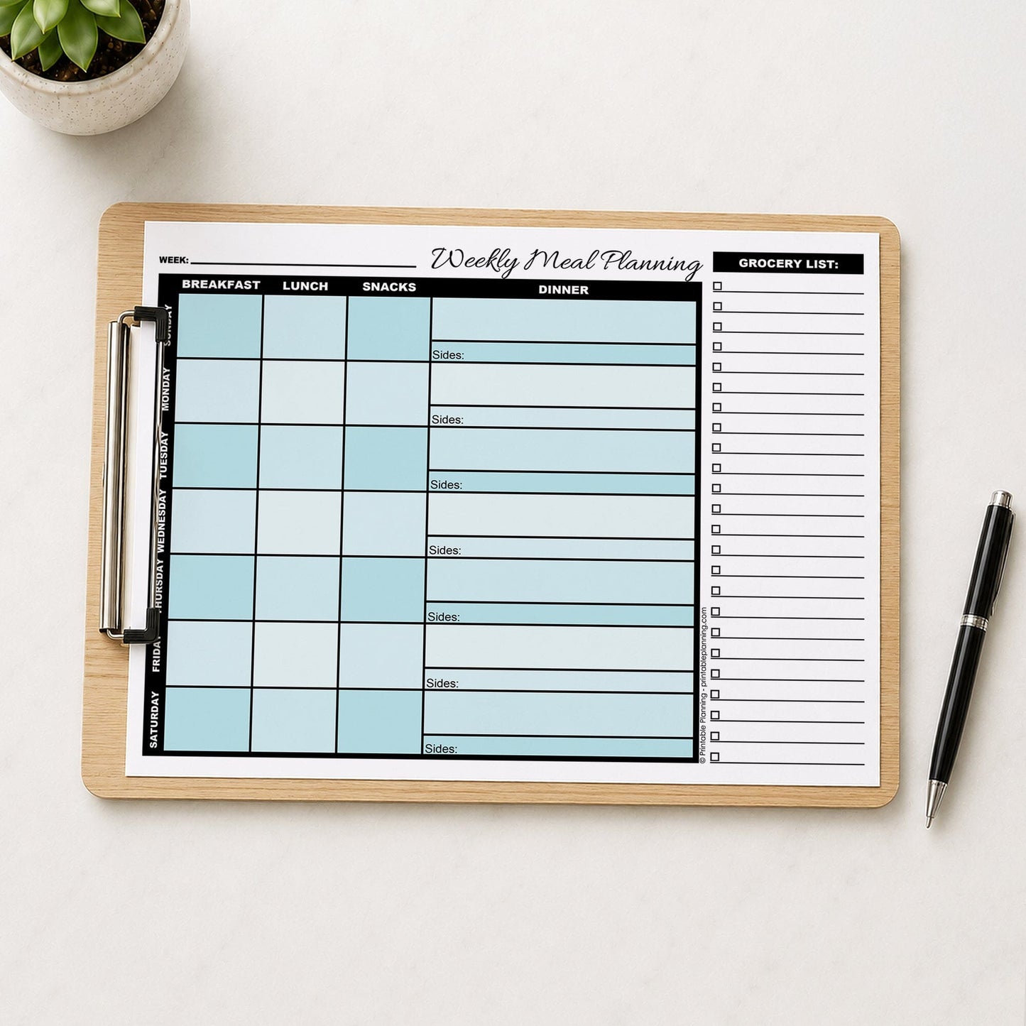A printed Turquoise Weekly Meal Planner with Grocery List (printable page) shown on a wooden clipboard on a table with a plant and a pen.