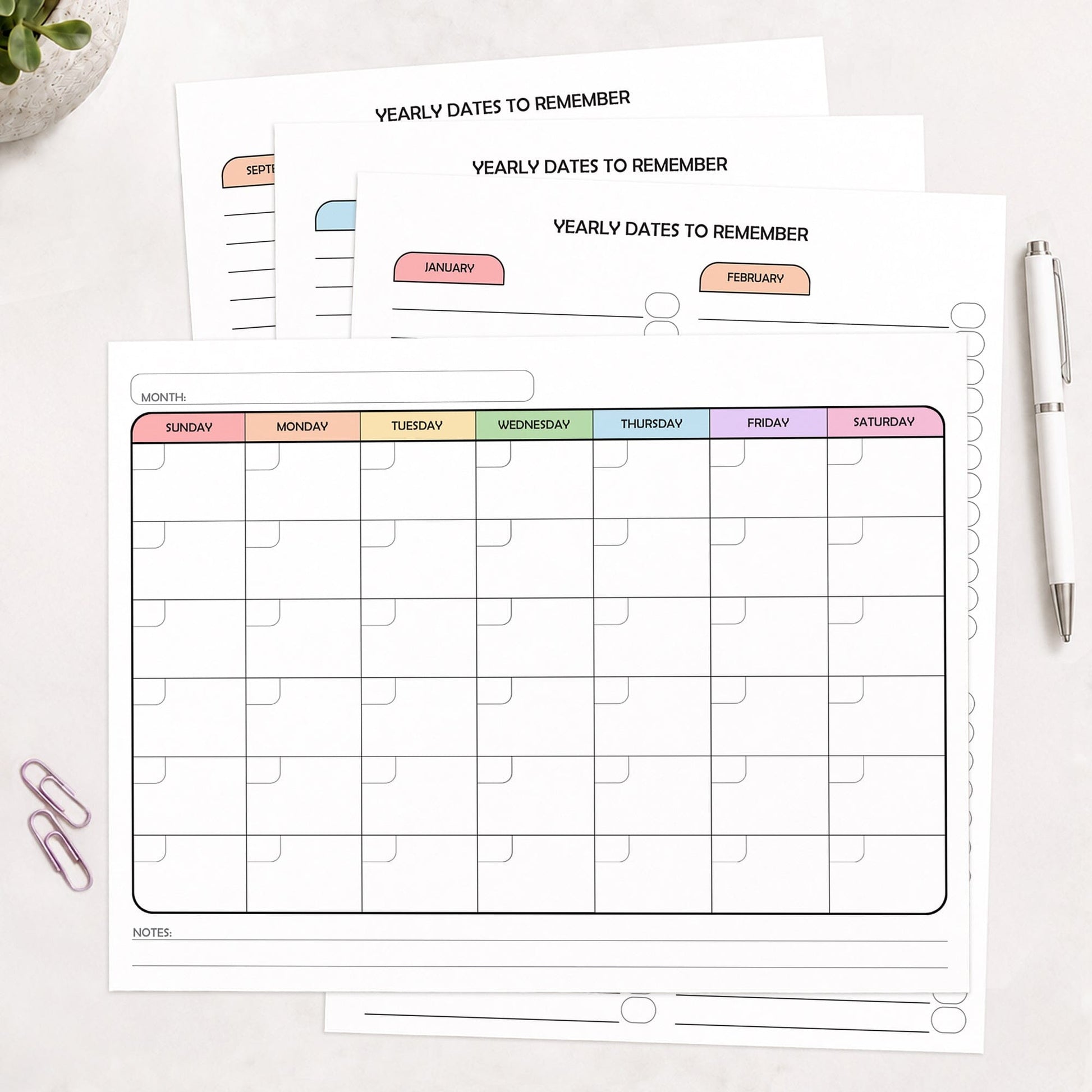 The 4 printed pages of the Undated Monthly Calendar & Important Dates Tracker Bundle shown on a desk with a plant, paperclips, and a pen.