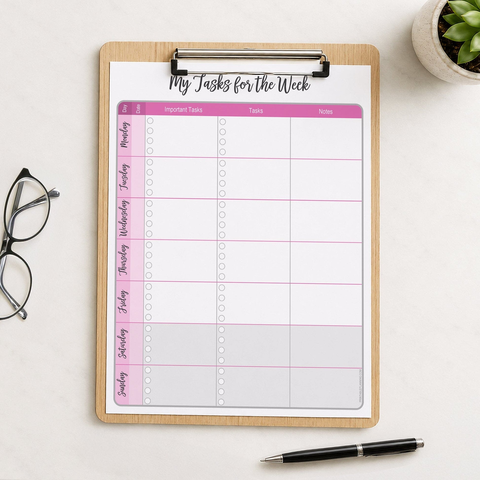 The printed Pink Weekly Task Planner with Priorities, Checklists & Notes (printable) page shown on a wooden clipboard on a table with a plant, glasses, and a pen.