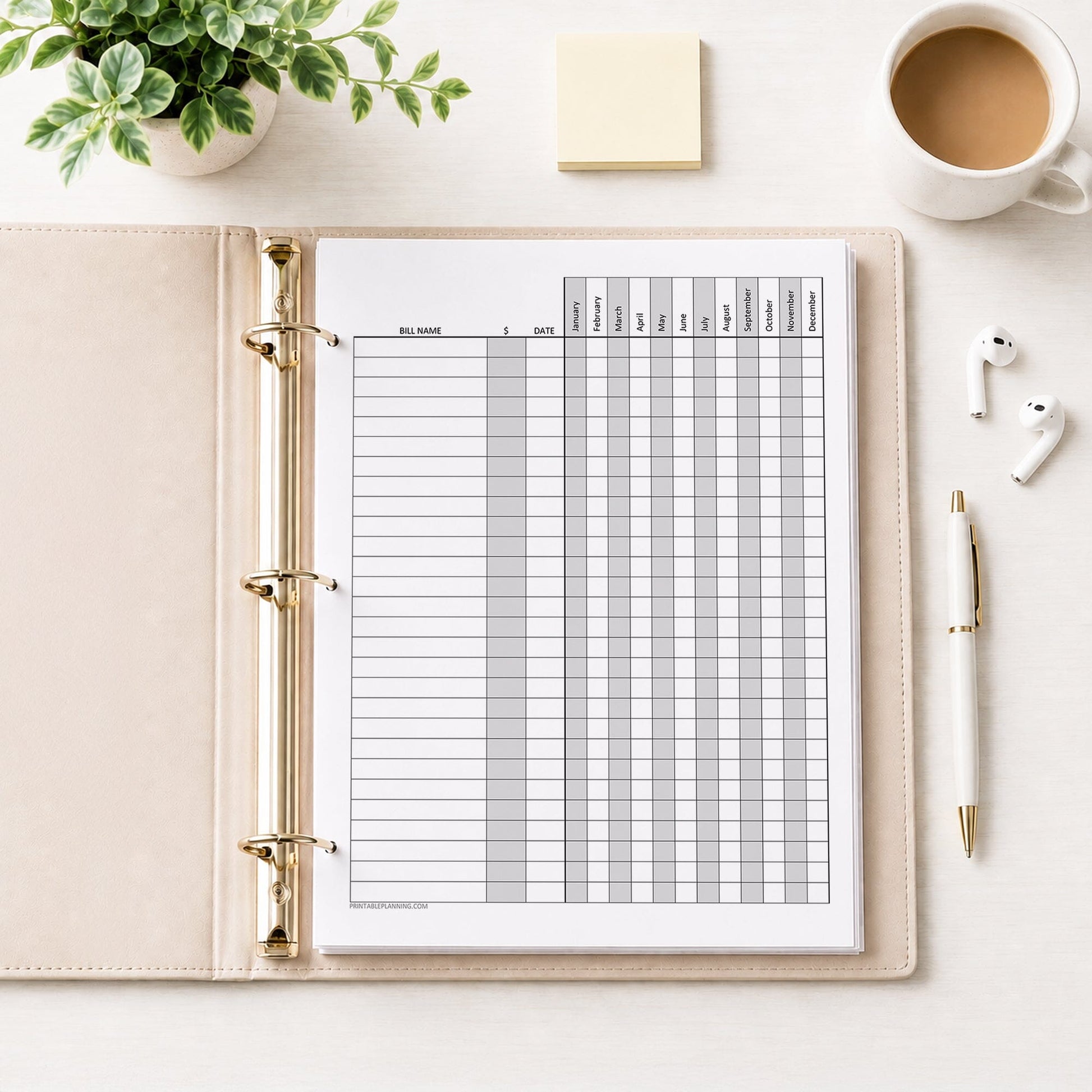 Yearly Bill Payment Tracker with Amount Column - Printable Annual Bill Checklist shown in a beige binder on a desk with earbuds, a plant, coffee, notes, and a pen.