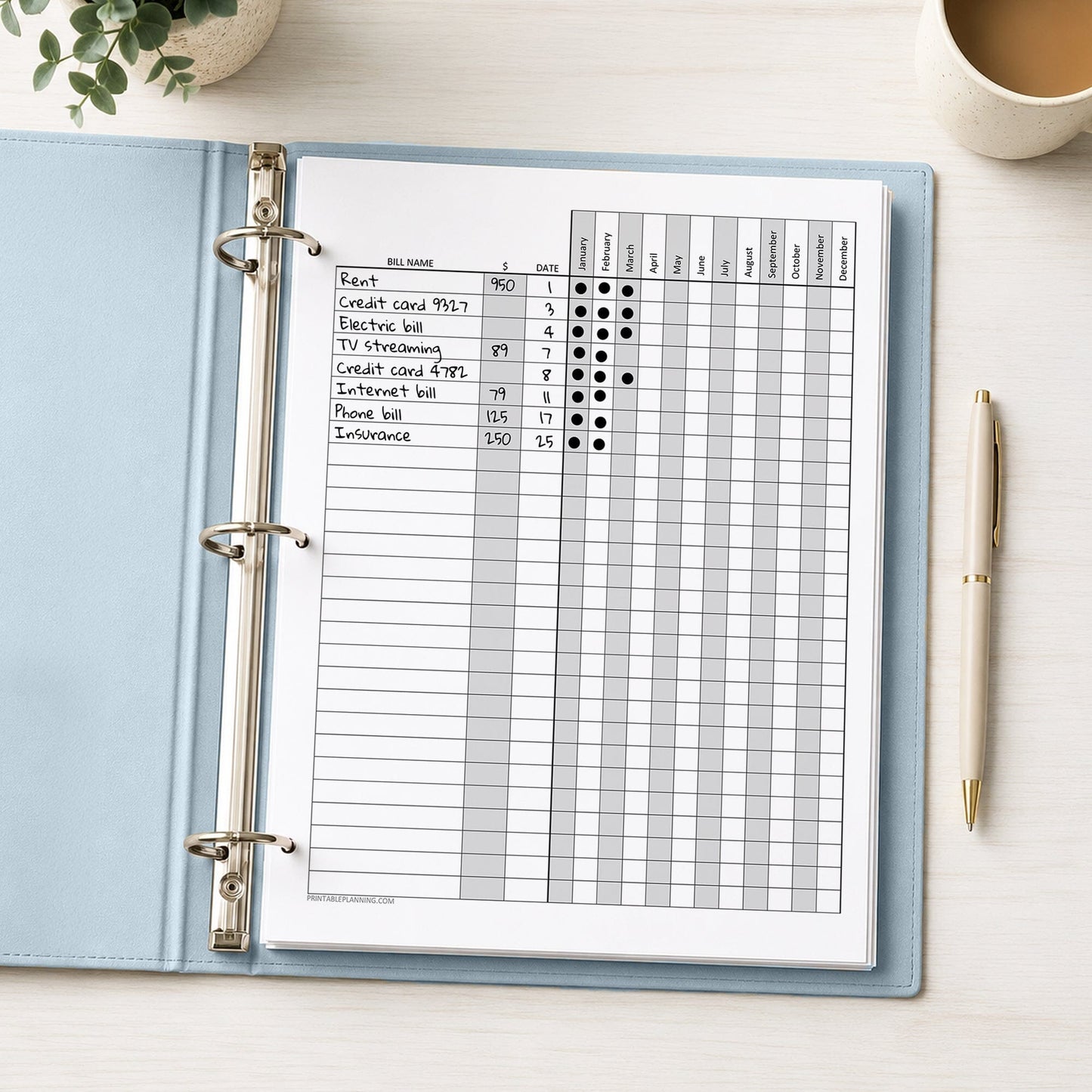 A printed Yearly Bill Payment Tracker with Amount Column (printable) page, shown with example writing, in a light blue binder on a desk with a plant, coffee, and a pen.