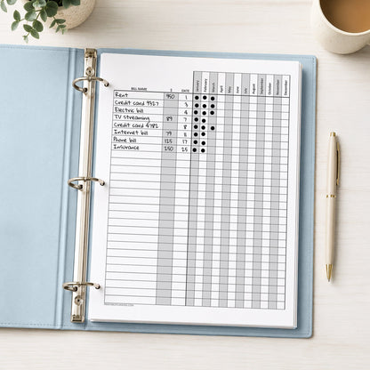 A printed Yearly Bill Payment Tracker with Amount Column (printable) page, shown with example writing, in a light blue binder on a desk with a plant, coffee, and a pen.