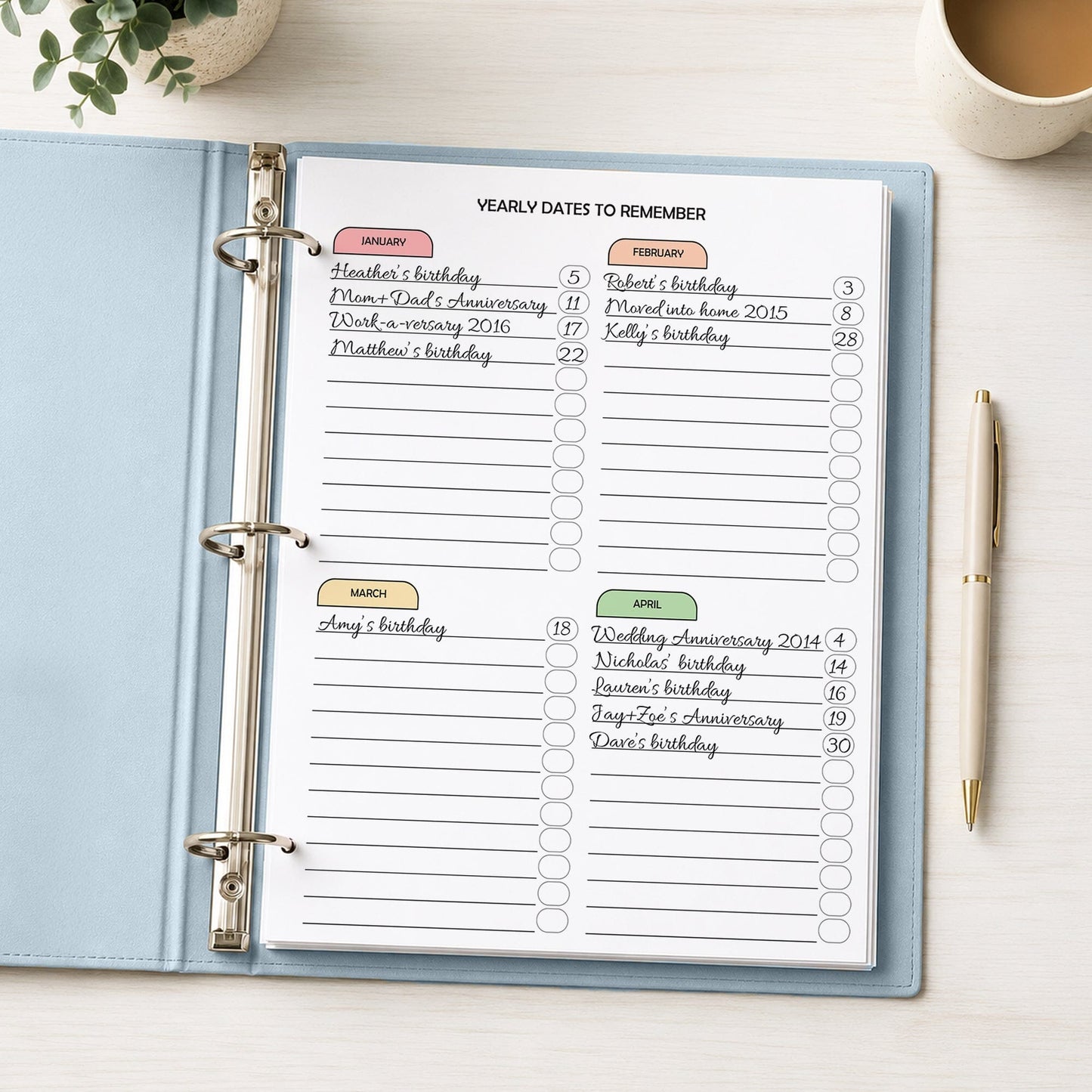 An open light blue ring binder planner showing printed monthly date reminder pages with handwritten entries, photographed on a white desk with a pen, plant, and coffee cup nearby.